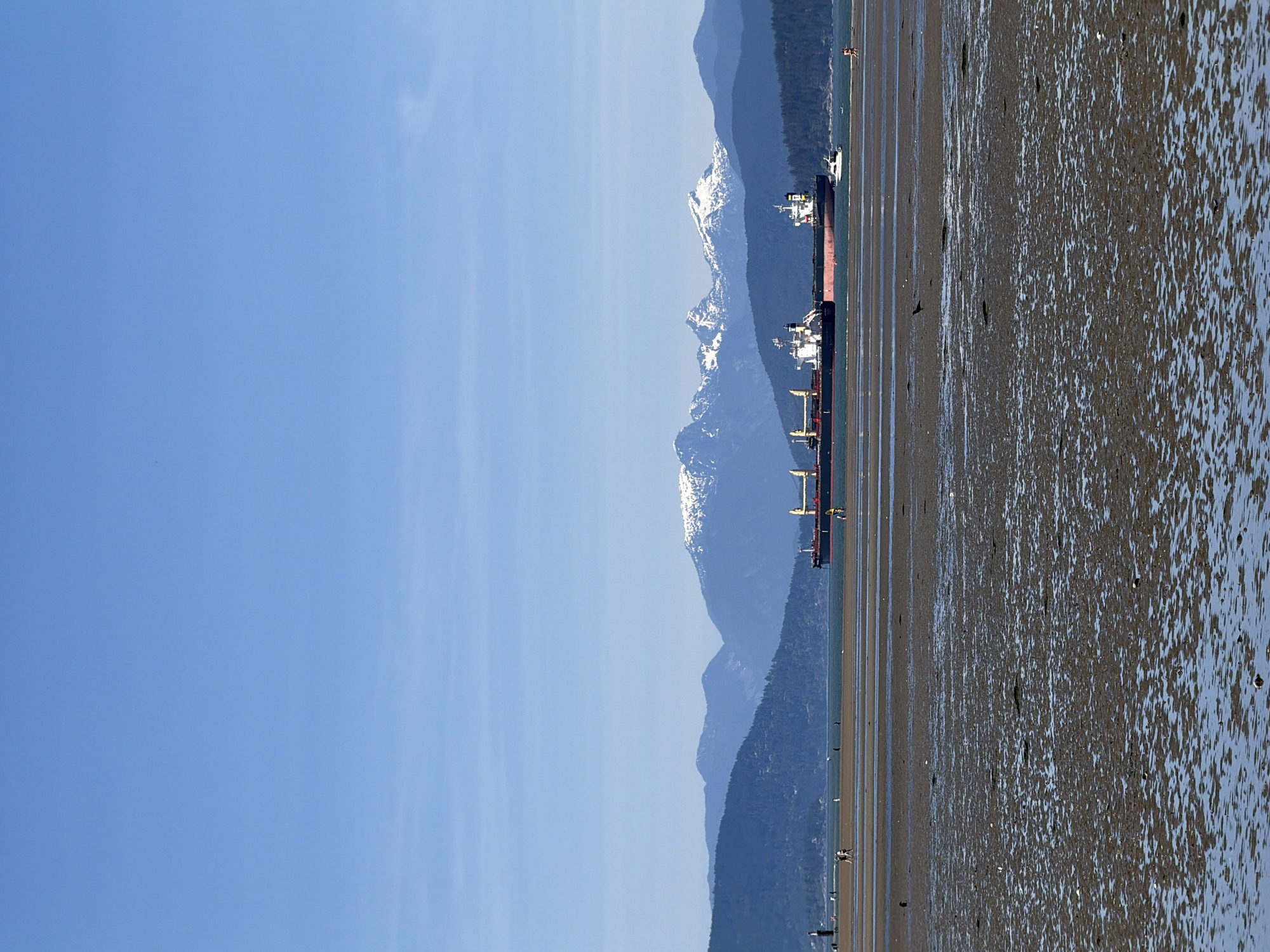 The image depicts a coastal scene in Vancouver, Canada, specifically from Jericho Beach facing north. Two large bulk carrier ships are anchored offshore in English Bay. In the background are the North Shore Mountains, featuring prominent, snow-capped peaks. The foreground shows the exposed tidal flats of Jericho Beach at low tide, with wet sand and pools of water reflecting the sky. A few people can be seen walking on the beach. The sky is clear and blue.