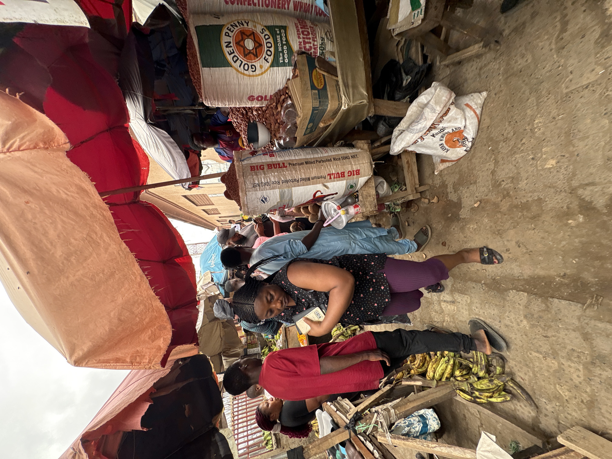 The image shows a crowded marketplace scene in Lugbe, Nigeria. Several people are gathered, interacting amidst various goods for sale. A woman in a floral top and purple leggings is at the center, holding a cell phone. Nearby, a man in a red shirt sits next to a pile of green bananas. Bags of "Golden Penny Foods" and "Big Bull Premium Milled Parboiled Rice" are stacked nearby. The marketplace is covered by a makeshift awning with various colors. The environment is bustling and typical of a local market.