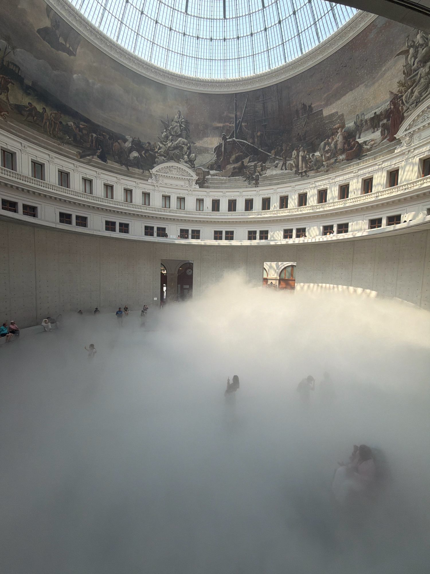 The image depicts the interior of the Bourse de Commerce in Paris, France, an historic circular building with a large glass dome. The central space of the building is filled with a dense, white mist or fog, obscuring much of the lower level and the people within it. Silhouettes of people are visible, some seated on benches along the outer wall, others standing or walking through the mist. Above the mist, the stark, concrete walls of the lower levels are visible, punctuated by windows. Higher still, a multi-tiered balcony with numerous windows encircles the space. The domed ceiling is constructed of glass panes and metal supports, allowing natural light to filter into the building. The upper walls of the interior are adorned with large, detailed historical murals depicting scenes of ships, people, and possibly battles. The overall impression is one of an immersive art installation or event creating an atmospheric and somewhat mysterious environment.