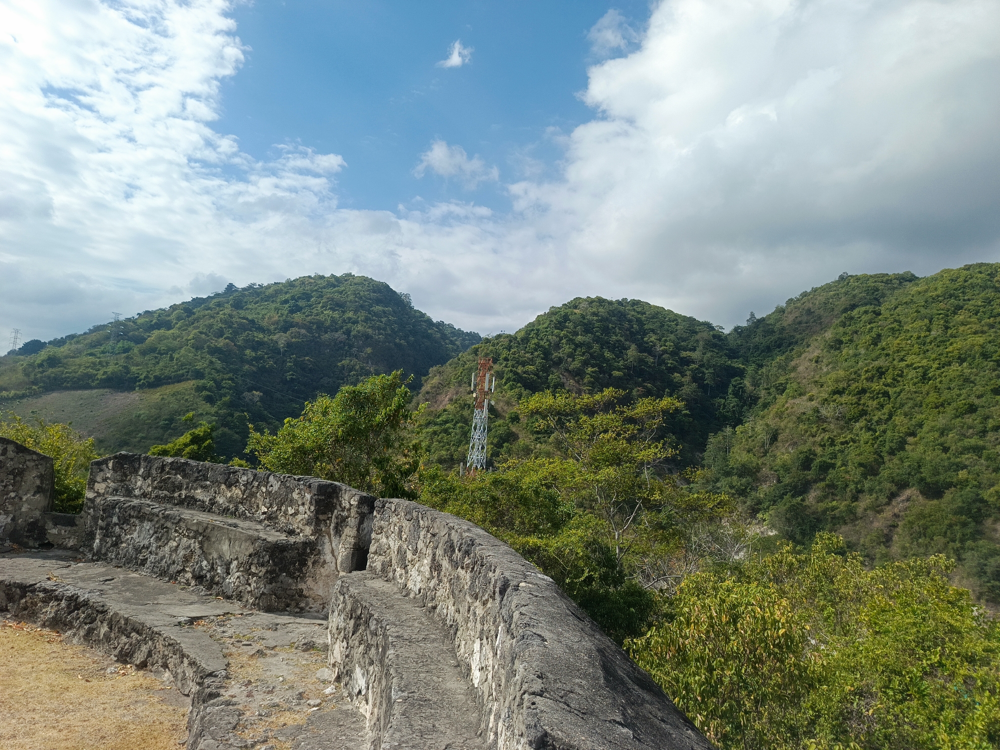 The image depicts a panoramic view of a lush, green mountainous landscape under a partially cloudy blue sky. In the foreground, there is a weathered stone structure with steps, resembling ancient ruins or fortifications, with dry, yellowish grass growing around its base. In the middle ground, dense green foliage covers rolling hills, and a tall, white metal telecommunications tower with orange accents stands prominently between two peaks. The background shows more distant, verdant mountains shrouded in a hazy atmosphere. The overall scene suggests a natural, undeveloped environment, possibly a historical site or a viewpoint overlooking a valley. This scene is consistent with the topography and vegetation found in the hilly areas surrounding Gorontalo, Indonesia, a region known for its natural beauty and historical remnants.