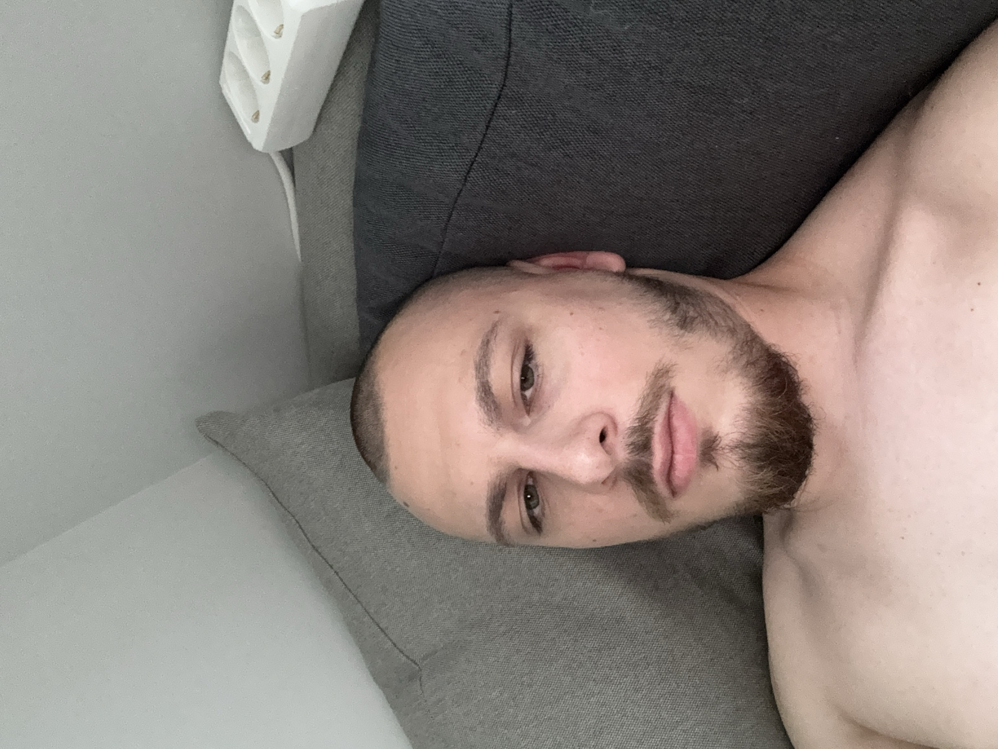 A man with a close-cropped haircut and a beard is lying on a gray pillow. His head is turned toward the viewer, and his upper torso is visible. He appears shirtless. He is lying next to a dark gray sofa. A white power strip is visible above the sofa. The location cannot be determined from the image. The city of Stara Zagora, Bulgaria cannot be determined from the image.