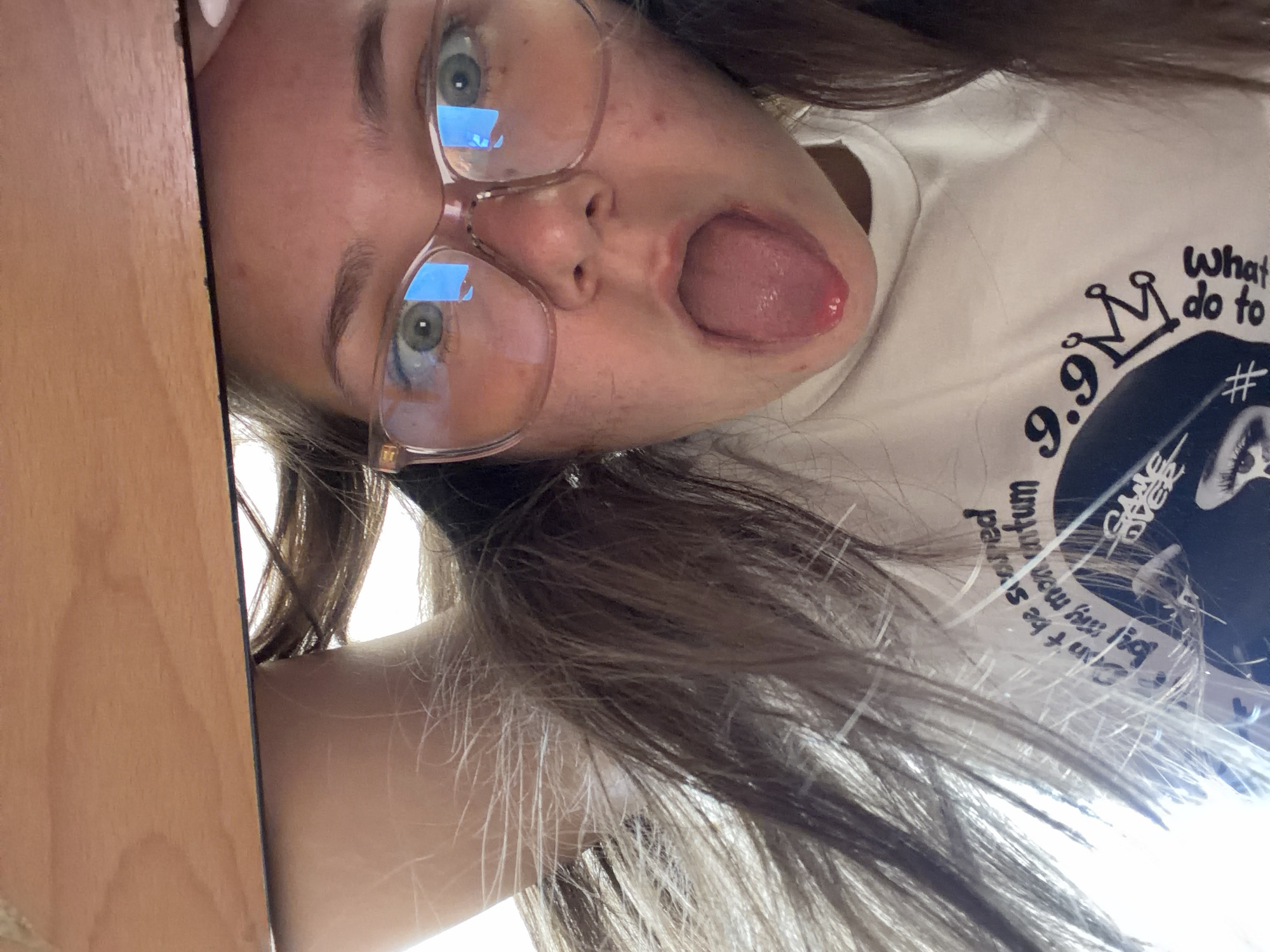 The image shows a person with fair skin, brown hair, and green eyes wearing clear-framed glasses. The person is sticking their tongue out towards the camera. They are wearing a white t-shirt with black and white graphics. A wooden surface is visible on the left side of the frame. The location is not evident from the image; there is no information indicating this image was taken in Veliko Tarnovo, Bulgaria.