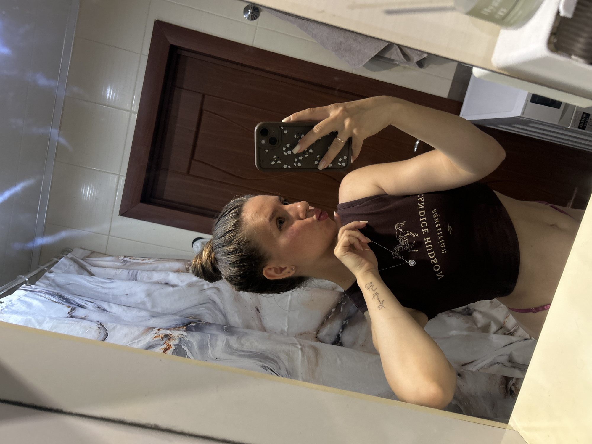 A young woman with her hair in a bun is taking a mirror selfie in a bathroom. She is wearing a dark crop top and pink underwear. She has a tattoo on her left arm and a necklace with a heart pendant. She is holding her phone in her right hand. The bathroom has a marble-patterned shower curtain and a wooden door. The image appears to be taken in a residential bathroom.