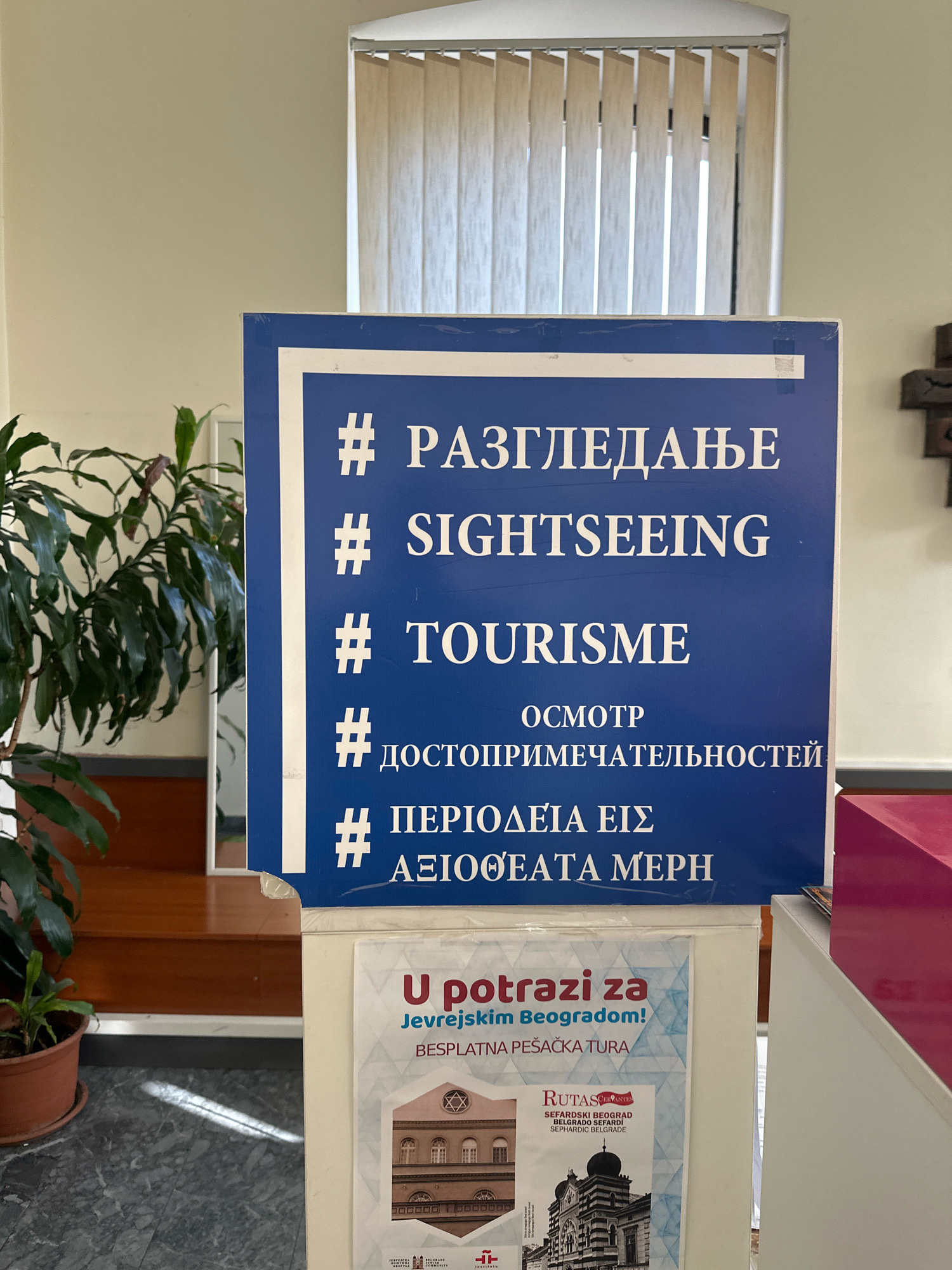 The image shows a blue sign with white text and hashtags. The text is in Cyrillic, Latin, and Greek alphabets, and appears to be a list of translations for the words "sightseeing" and "tourism." Below the blue sign is a poster advertising a free walking tour of Jewish Belgrade. The poster features images of Jewish landmarks in the city and text in Serbian and English. The overall impression is of a tourist information sign or brochure. The scene is indoors, possibly in a public building or tourist center, given the presence of the sign and poster. There are no people visible in the image.