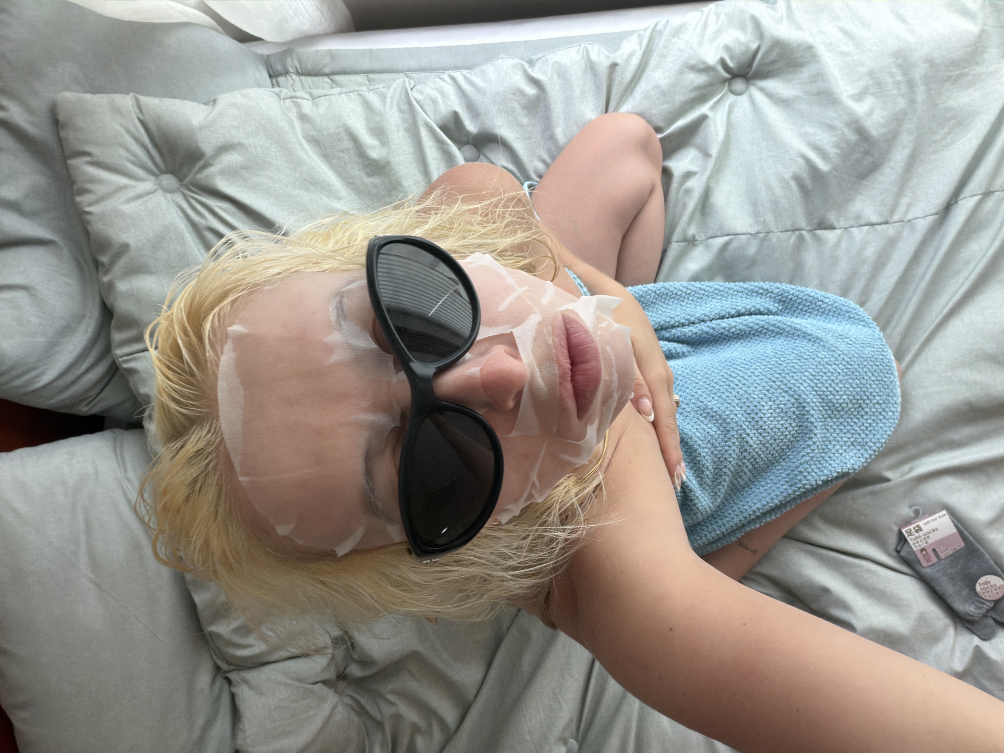 A woman lies on a bed, her head resting on a light gray, button-tufted pillow. She is wearing a sheet mask on her face and black sunglasses over the mask. Her blonde hair is spread out around her head. She is partially covered by a light blue towel. A package of socks is visible on the bed near her legs. The location is not discernible from the visual information provided. The city of Seoul, South Korea, is not identifiable in this picture.