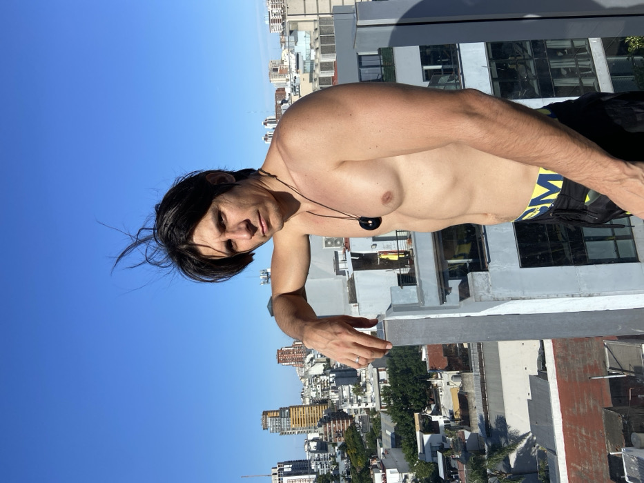 A shirtless man with dark hair stands on a rooftop, with a city skyline in the background. He wears a black pendant necklace and has dark shorts, possibly swim trunks, with yellow accents. He is leaning on the ledge of the roof, partially obscuring the cityscape behind him. The sky is clear and blue.