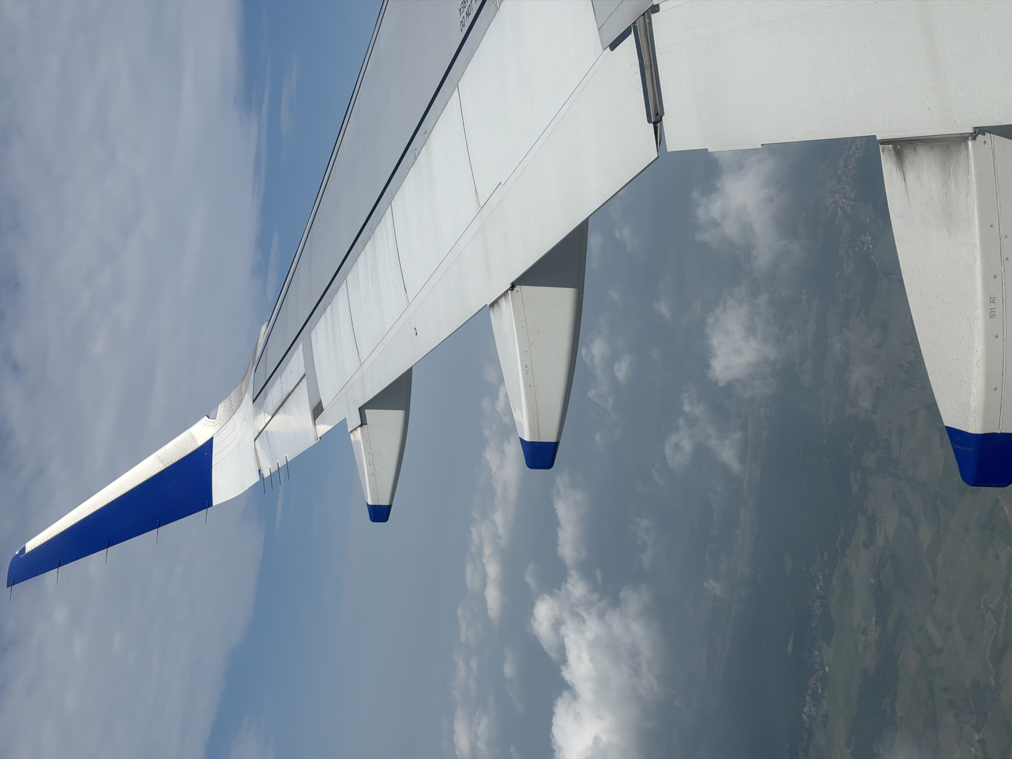 The image captures an aerial view from an airplane window, focusing on the airplane's wing and the landscape below. The wing, predominantly white with blue accents at its tips, extends diagonally across the frame. The sky is a mix of blue and white clouds, some appearing dense and gray, suggesting overcast conditions at lower altitudes. Below the clouds, a patchwork of green fields and a small village or town, likely Mohan, India, is visible. The village appears as a cluster of buildings nestled within the rural landscape, with roads or paths subtly discernible. The overall scene depicts a typical view during an airplane flight over a populated rural area.