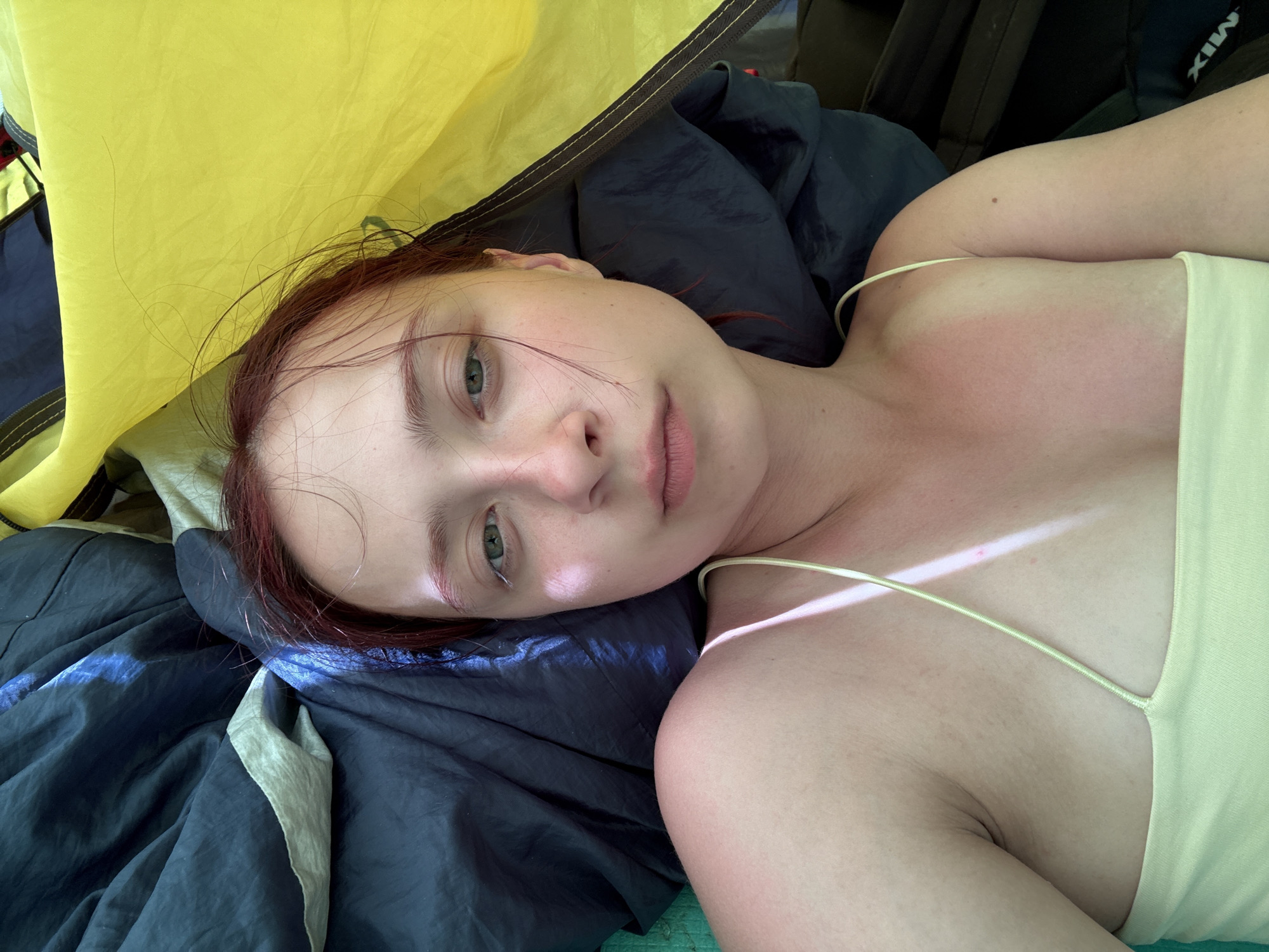 A young woman with light skin and red hair lies on a dark blue and gray wrinkled fabric inside a yellow tent. She wears a light yellow tank top with thin straps. Her shoulders are slightly reddened, possibly from sunburn. Her face is angled toward the camera, and she has a neutral expression. A backpack is visible in the upper right corner. The location is unknown, and there is no evidence to indicate the location is in Khvalynsk, Russia.