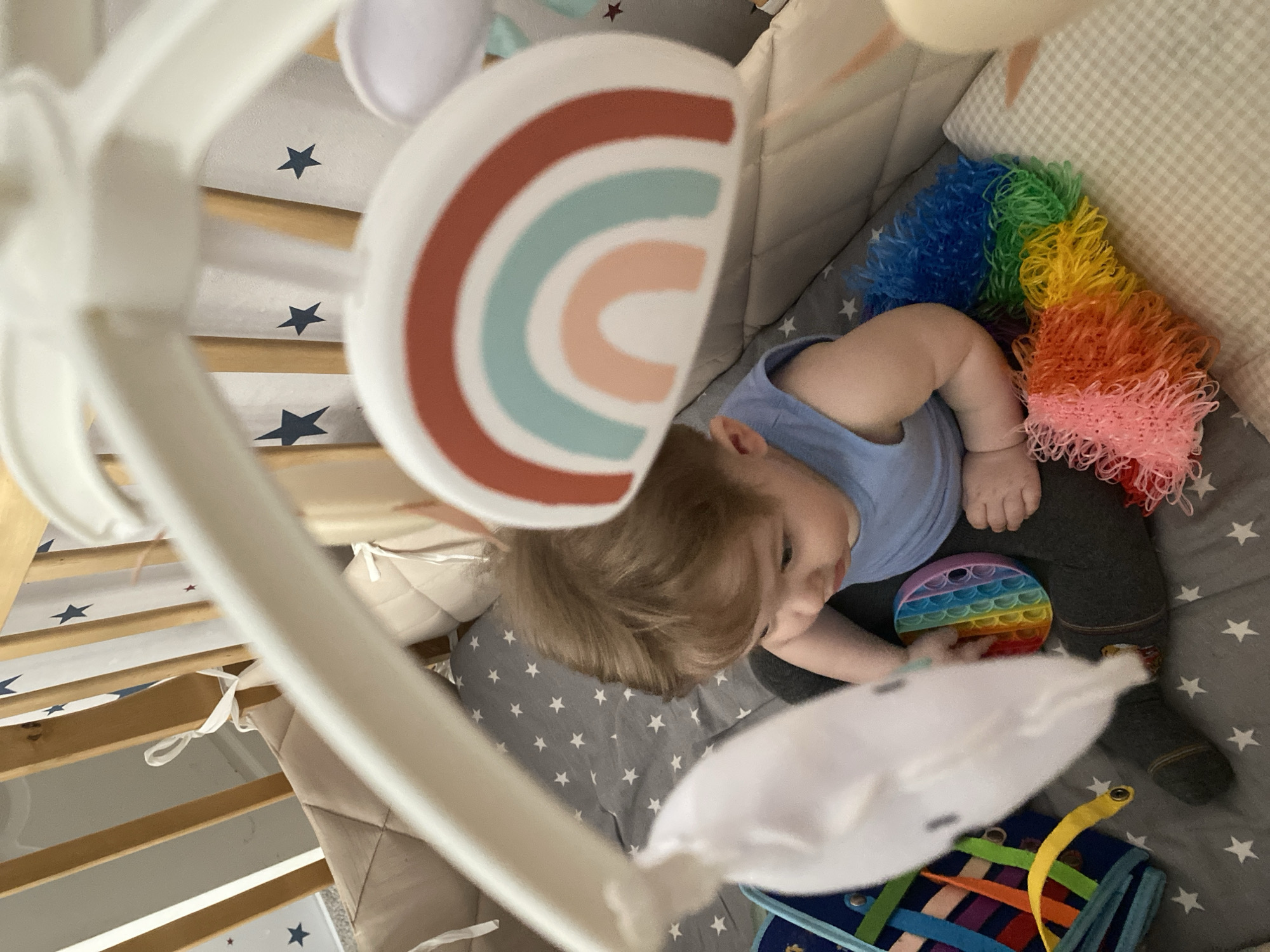 A baby lies in a crib in Votkinsk, Russia. The baby wears a blue shirt and dark pants and holds a rainbow-colored "pop it" fidget toy. A rainbow-colored toy rests at the head of the crib, and a blue fabric toy is at the foot of the crib. The crib is lined with padded bumpers, and a baby mobile hangs above. The crib sheet is gray with white stars. The crib is constructed of wood with white painted sections.