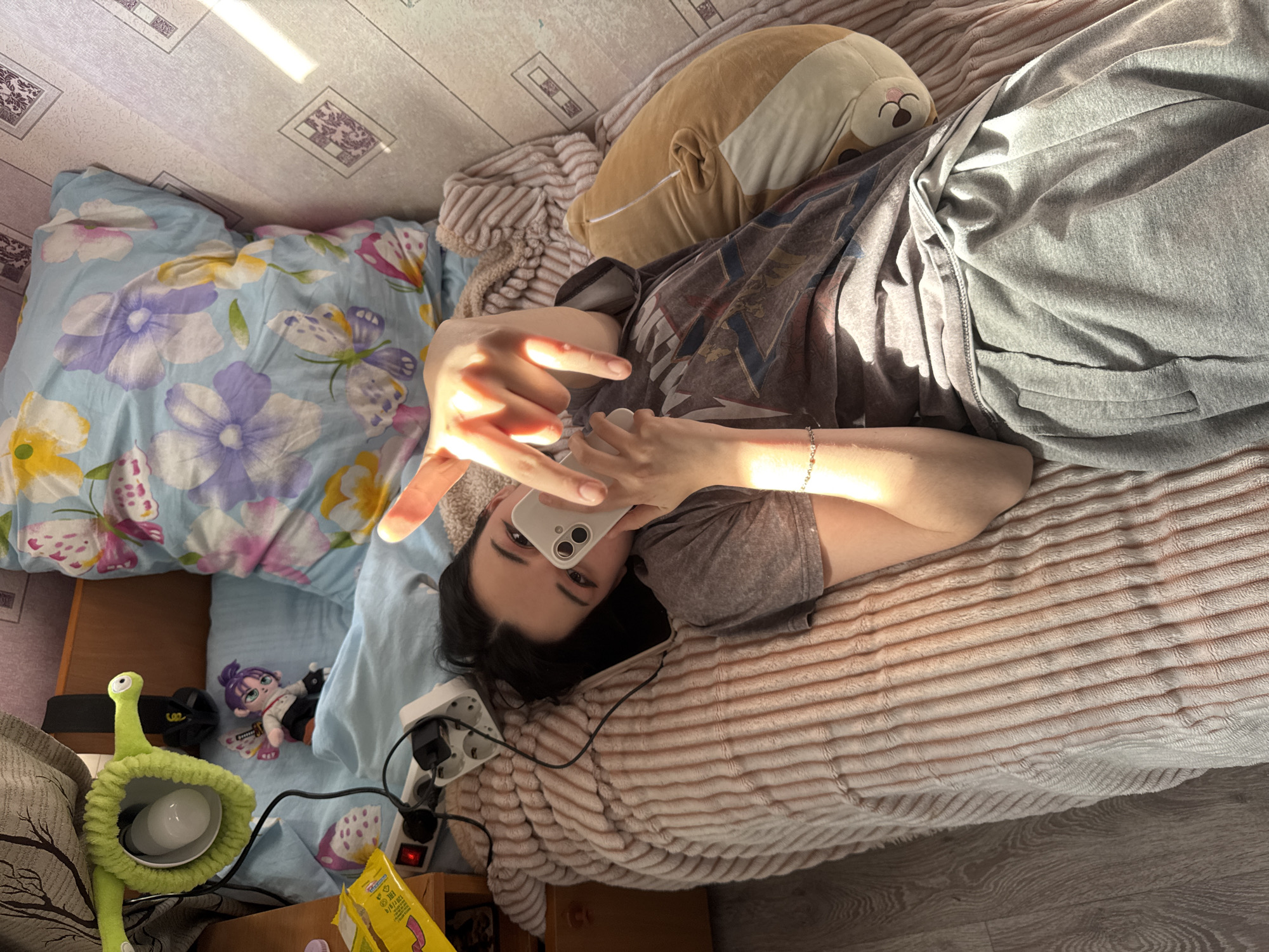 The image presents an overhead view of a person lying on a bed or daybed in an indoor setting, likely a bedroom, under bright daytime sunlight. The subject, with dark hair, wears a grey graphic t-shirt and grey sweatpants. They are holding a white smartphone with two camera lenses, looking directly at the camera, suggesting they are taking a selfie or engaged in a video call. Their left hand is raised, making a gesture (resembling a "rock on" sign), and is strongly illuminated by sunlight.
The bed features several textiles: a light blue duvet patterned with purple and yellow flowers and butterflies, a plain light blue sheet, and a fuzzy beige or light brown striped blanket. Resting near the person's head is a large, light brown plush toy, possibly a dog or corgi, wearing a small white face mask. A smaller, smiling corgi-like plush toy and a small doll with purple hair and black and white attire are also visible on the bed.
To the left, on a wooden surface, is a distinctive green plush lamp with a flexible body, resembling an alien or snail, holding a white light bulb. A white power strip with a red switch and various connected chargers/cables is positioned near the person's right. Partially visible yellow packaging with Russian text, including "Состав" (Ingredients), "Энергетическая ценность" (Nutritional value), and "Масса нетто: 60 г" (Net weight: 60 g), indicates a snack item, likely chips. The floor has a light-colored patterned linoleum or laminate. The bright sunlight suggests a sunny day. The location context is Perm, Russia.