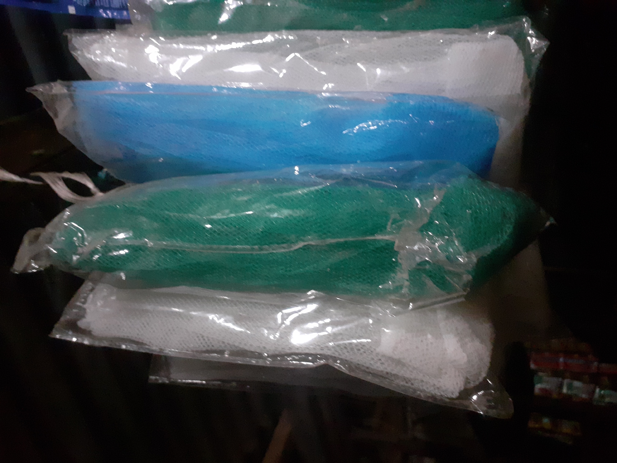 This image, taken in Jalingo, Nigeria, shows a close-up view of several mesh loofahs hanging together. The loofahs are packaged in clear plastic and are of various colors, including white, green, and blue. They are arranged in a line, with the white loofahs on the left and right, and green and blue loofahs in the middle. The background is dark and out of focus, with some indistinct shapes that might be shelves or other items in a store. The lighting suggests an indoor setting, possibly a market stall or a small shop.