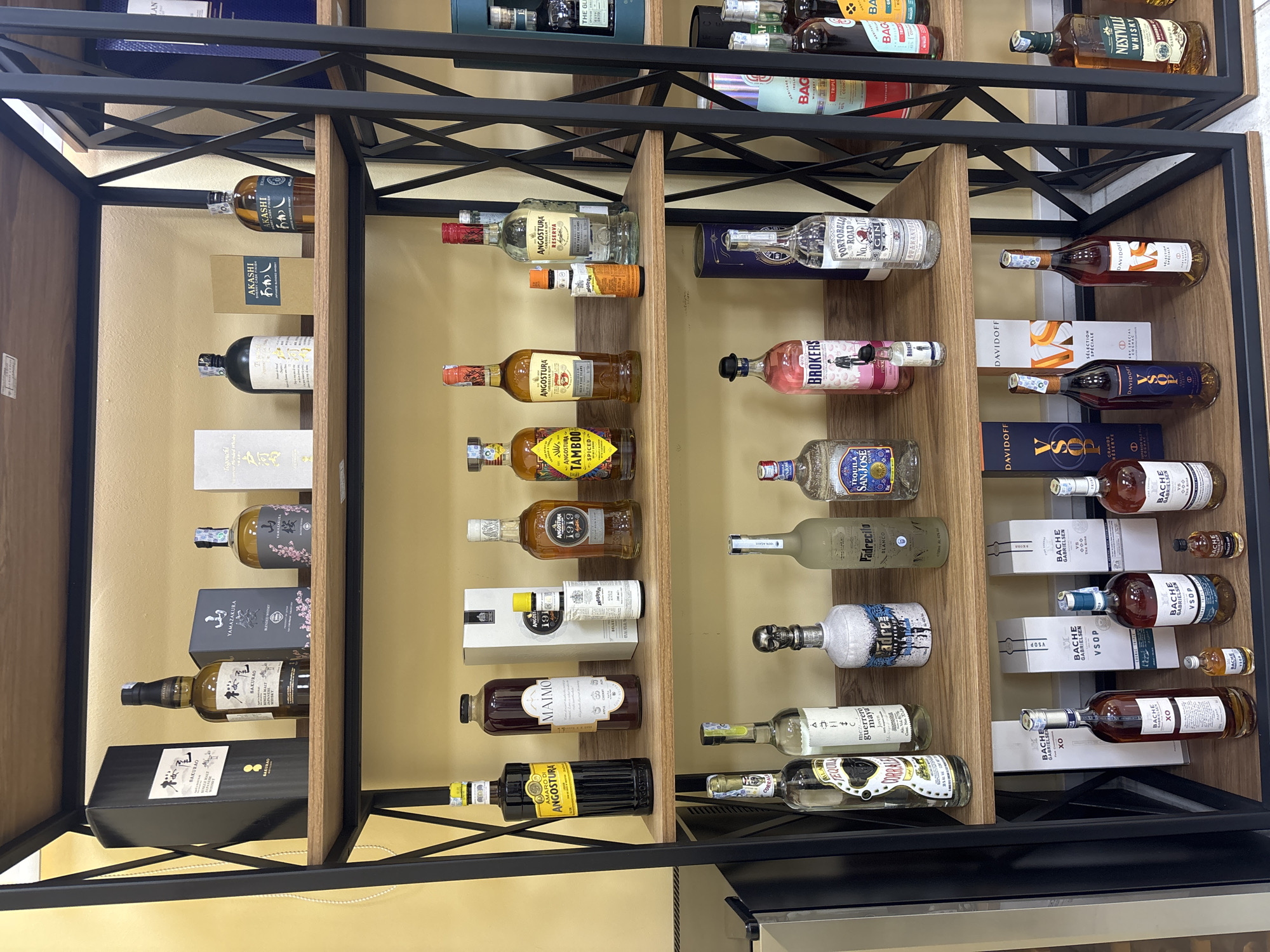 This is a photograph of shelves of various alcoholic beverages, possibly in a store or a bar. The drinks include different types of whiskey, gin, tequila, and liqueurs. Brand names and styles visible include Angostura, Akashi, Yamazaki, and Davidoff. The shelving unit is made of a light-colored wood, with a black metal frame. The location is likely a retail setting, but the exact location in Tbilisi, Georgia is not possible to determine.