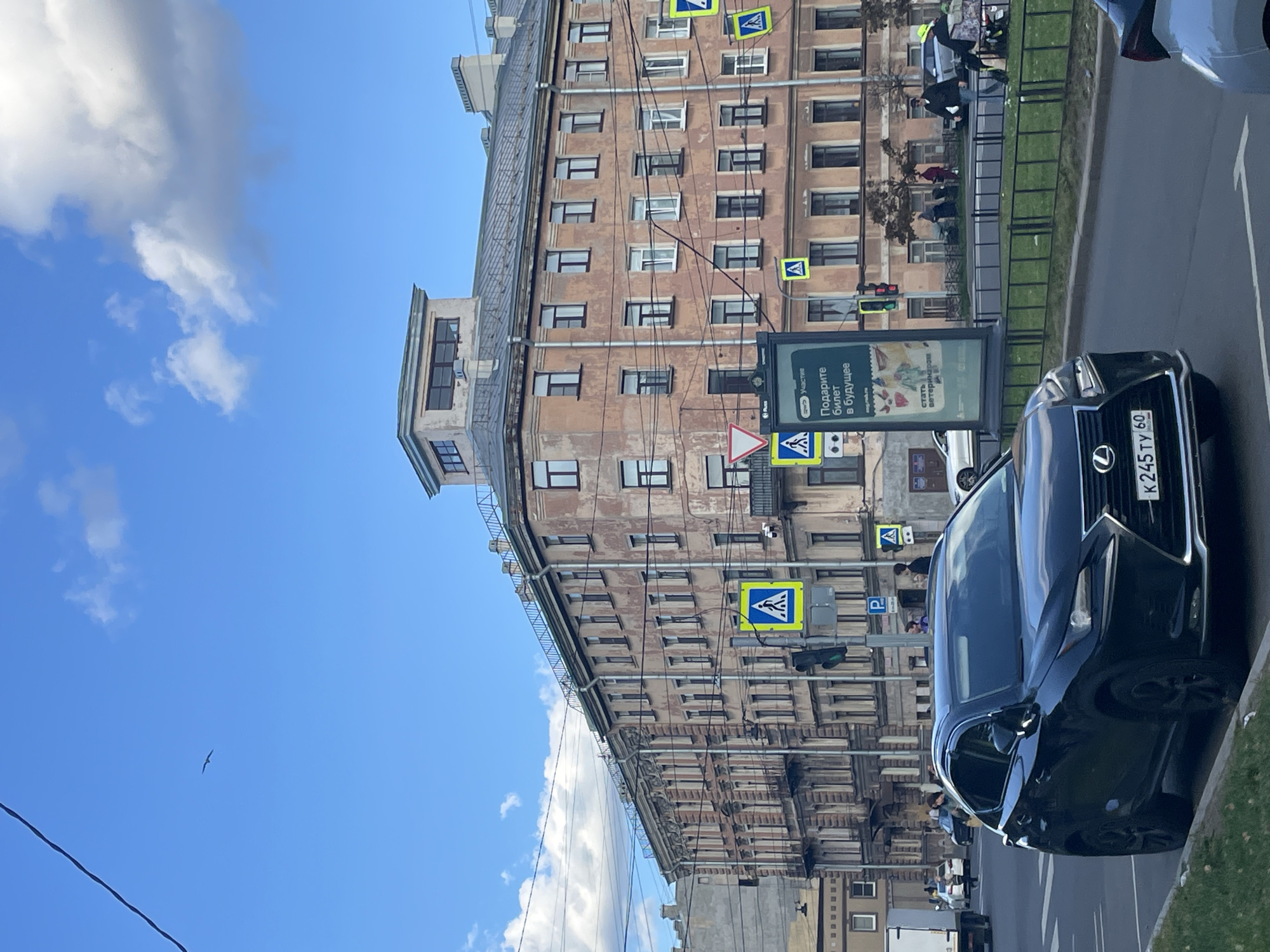 The image depicts a street scene in Saint Petersburg, Russia, likely during the daytime under a partly cloudy sky. In the foreground, a black Lexus SUV is parked or stopped by the curb. Behind the SUV, a street with multiple lanes of traffic is visible, with other vehicles and pedestrians present.
Dominating the background is a large, multi-story building with an aged, textured facade, characterized by rows of windows and balconies. The architecture suggests a pre-Revolutionary or early Soviet era style. Across the street, a variety of street signs are visible, including pedestrian crossing signs and a parking sign. A large advertising display stands near the building.
Several people are seen walking on the sidewalk and near the street, some with strollers. Power lines crisscross the sky, and a distant bird is visible against the blue sky. The overall impression is of an urban environment with typical city elements like traffic, buildings, and pedestrians.