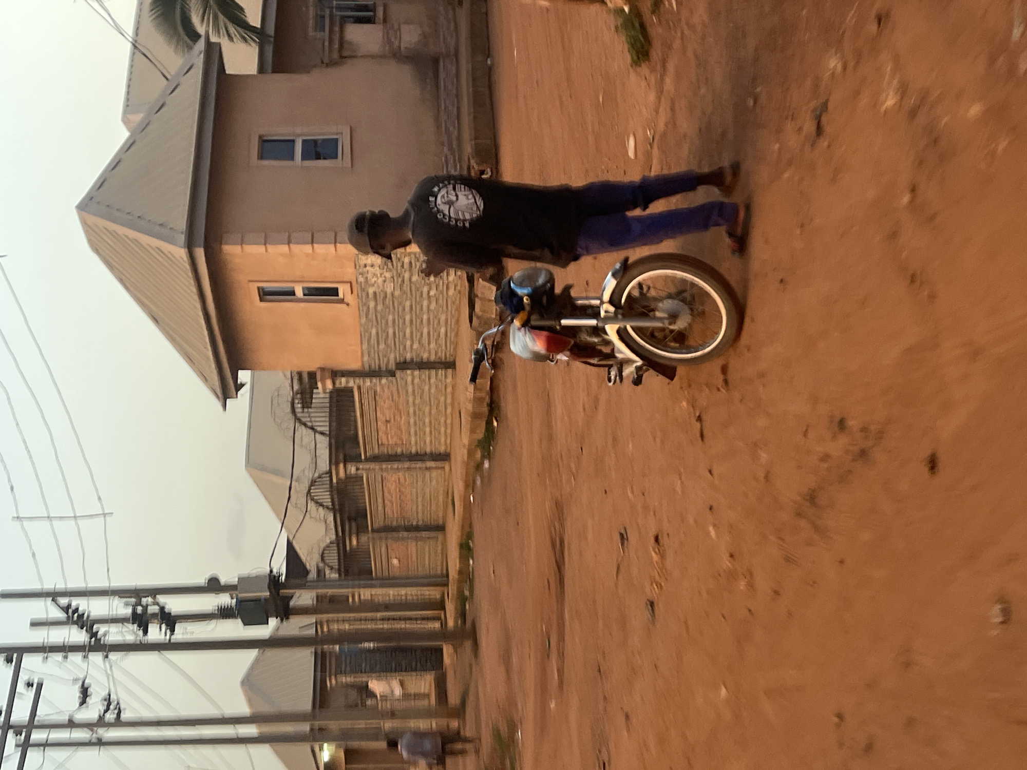 A medium shot shows a male figure with a black shirt, long purple pants and flip flops next to a motorbike on a dirt road. There is residential housing on the left and overhead power lines. The location is likely in Abuja, Nigeria.