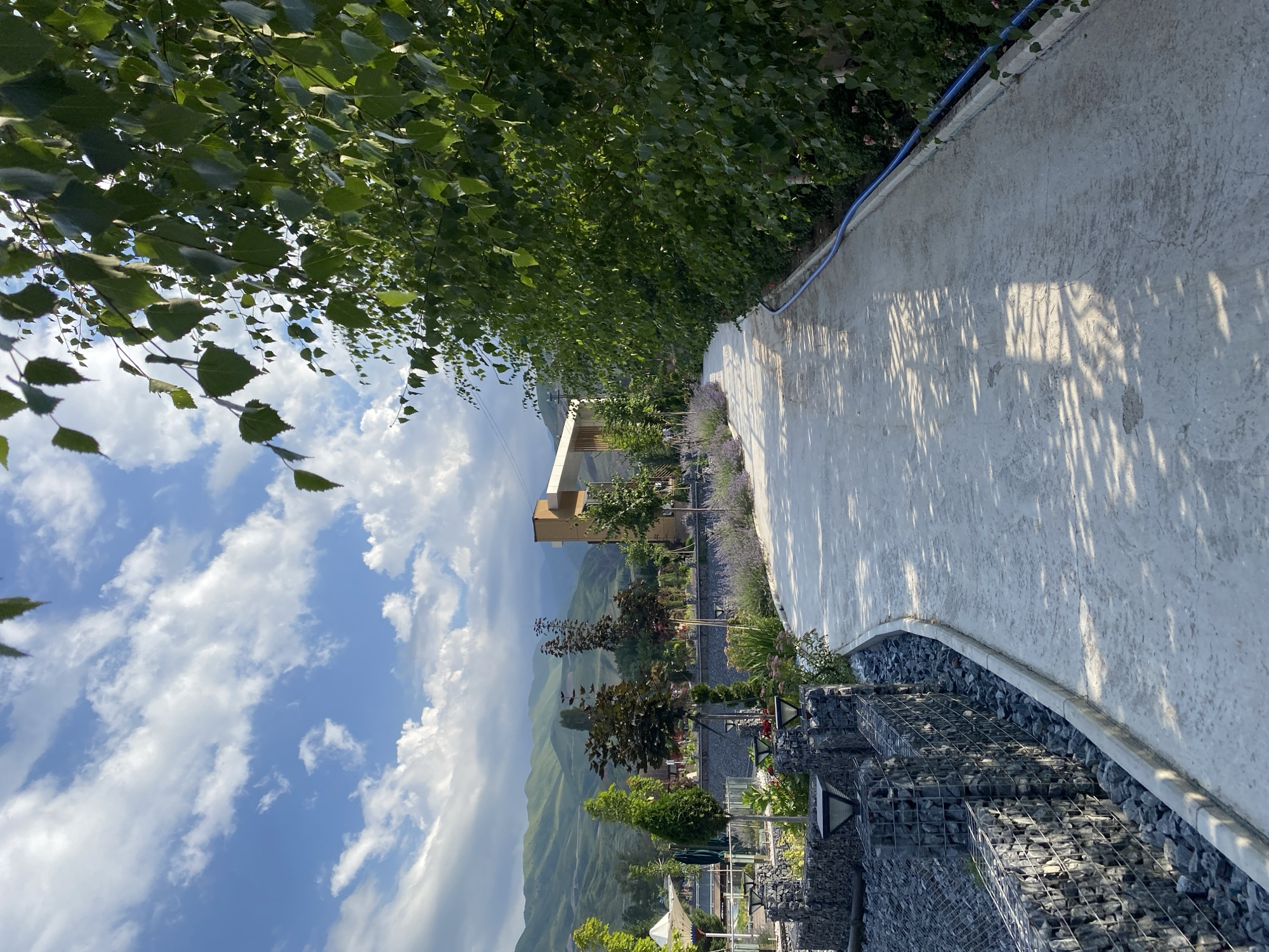 The image depicts a paved pathway curving through a landscaped area with mountains in the background. The pathway is lined on the left by gabion walls filled with dark stones and a low border, with trees and decorative plants behind them. On the right, a dense line of green trees and foliage borders the pathway, with a blue hose visible along the edge. In the distance, a modern building with wooden accents stands amidst more greenery and trees. The sky is blue with scattered white clouds, and sunlight casts shadows on the pathway. The scene appears to be a public park or garden area in Arashan, Kyrgyzstan. There are no people or animals visible in the image.