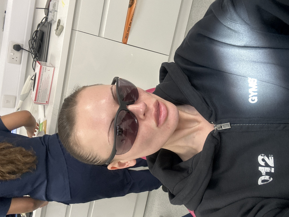 A female subject is depicted in a seated position in a clinical environment in Tunbridge Wells, United Kingdom, directly facing the camera. She wears black framed, dark-lensed protective glasses that obscure her eyes and a black zippered hoodie with "012" printed on the left chest and "GYMSHARK" on the right shoulder area. Her hair is pulled back, revealing a high hairline, and her facial features include prominent cheekbones and full lips. In the background, another individual, partially visible from the waist up, stands with her back to the camera. This individual wears dark blue medical scrubs and has long, wavy dark brown hair. She is positioned at a white countertop. On the countertop, a white box labeled "POWDER-FREE NITRILE EXAMINATION GLOVES" (Size L) is visible, alongside a white medical device, an electrical outlet with a plugged-in cable, a laptop, small clear containers, and a metallic instrument. White cabinetry lines the background, with a yellow/orange strip of tape partially visible on a cabinet door to the right, bearing the fragmented text "Will not be accessib..." and "DP".