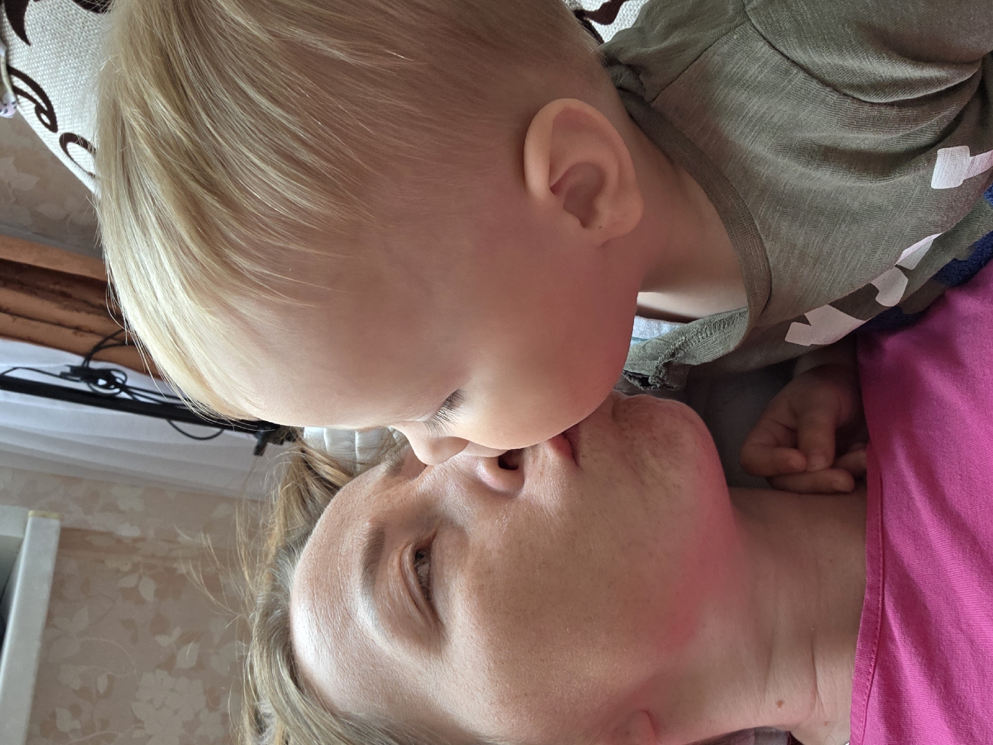 This image captures a close-up, intimate scene within an indoor domestic setting. While the user specifies the city of Chaikovsky, Russia, the visual content of the image does not provide any specific identifiers to corroborate this geographical location.
The primary subjects are an adult and a young child engaged in an affectionate interaction. The adult, appearing to be female, is positioned on her back, her head resting out of frame. She has light brown hair, visible wrinkles on her forehead and around her left eye, and scattered pigmentation marks on her cheeks. Her lips are distinctly pursed, and her left eye is slightly open. A small, dark mole is visible on her neck. She is wearing a bright pink top with a simple rounded neckline.
The child, a toddler with very light blonde, fine hair, is positioned on the right side of the frame, facing and leaning into the adult. The child's right eye is closed, and its face, specifically the mouth/cheek area, is pressed against the adult's pursed lips, indicating a kiss or nuzzle. A small, round ear is visible on the child's right side. The child is wearing a drab olive-green t-shirt with white printed letters visible on the left shoulder and chest area. A small, closed fist belonging to the child is visible near the adult's neck/collarbone area.
The background reveals a patterned wallpaper on the left, featuring light floral designs against a light base color. Above this, a white horizontal surface, possibly a shelf or windowsill, is present. On the right side, a sheer white curtain is partially visible, along with a dark vertical support structure and some dark cables. A light-colored cushion or pillow with a dark brown, ornate scroll pattern is visible behind the child's head. The lighting appears soft and consistent with an indoor environment.