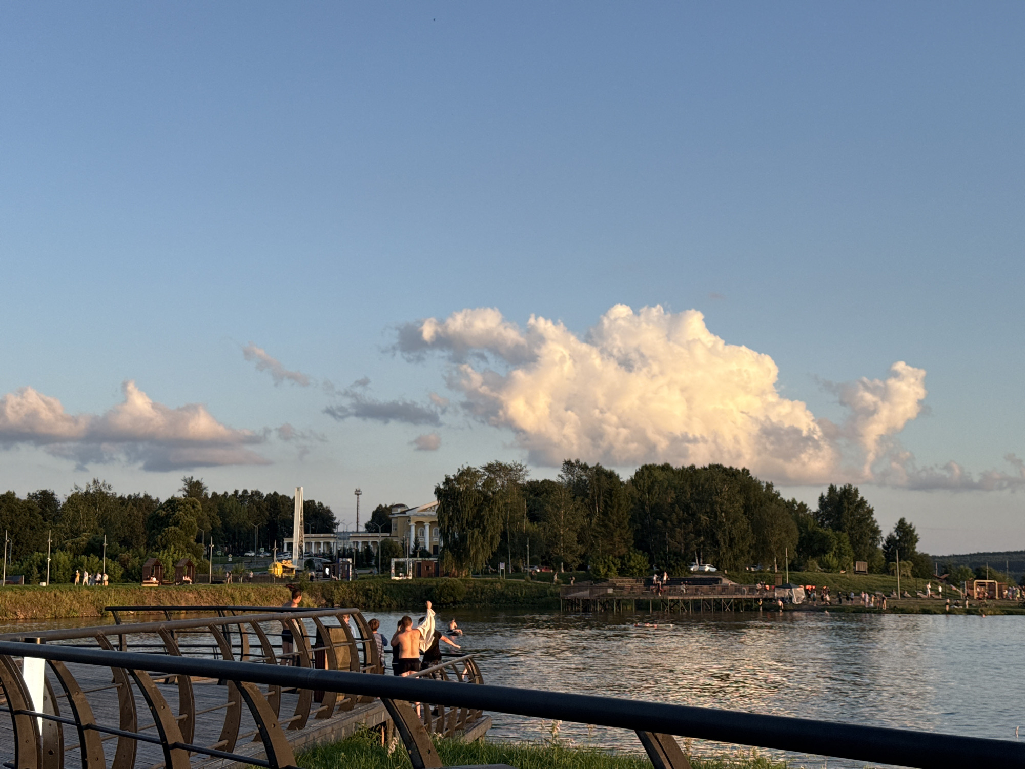 The image shows a waterfront scene in the city of Lysva, Russia, likely near the embankment. In the foreground, a dark metal railing with a curved design separates a wooden walkway from a body of water. Several people are visible on the walkway, some appearing to be in swimwear, suggesting they are preparing to enter or have just exited the water. The water itself reflects the golden light of the setting sun, casting a warm glow.
Across the water, a treeline forms the background, with a large, ornate white building with columns visible amongst the trees. Further along the embankment, more trees and structures are present, including what appears to be a pier or boardwalk extending into the water with people on it. A tall, white, obelisk-like monument stands out against the sky. The sky above is a clear, pale blue, with large, fluffy white clouds illuminated by the sun, indicating it is late afternoon or early evening. Scattered throughout the scene are various trees, some appearing as dense green foliage, others as individual specimens. There are also indications of public amenities such as gazebos or shelters along the grassy bank. The overall impression is of a public recreational area being enjoyed during pleasant weather.