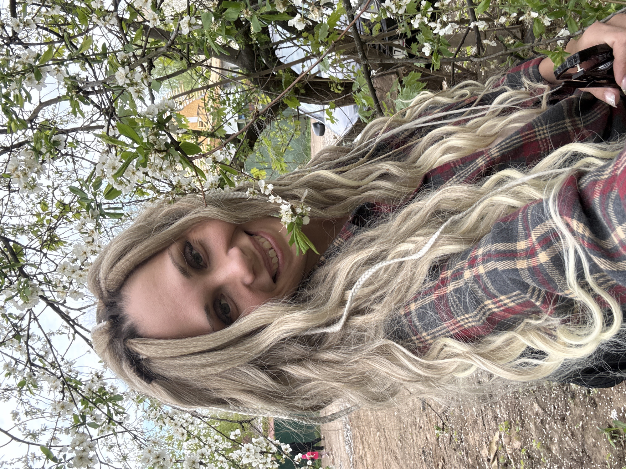The image shows a woman with blonde hair and a plaid shirt standing in what appears to be a wooded area in Rusanovo, Russia. She is smiling and looking at the camera. There are blooming trees with white flowers surrounding her. She is holding sunglasses. The ground appears to be covered with dirt and some vegetation.