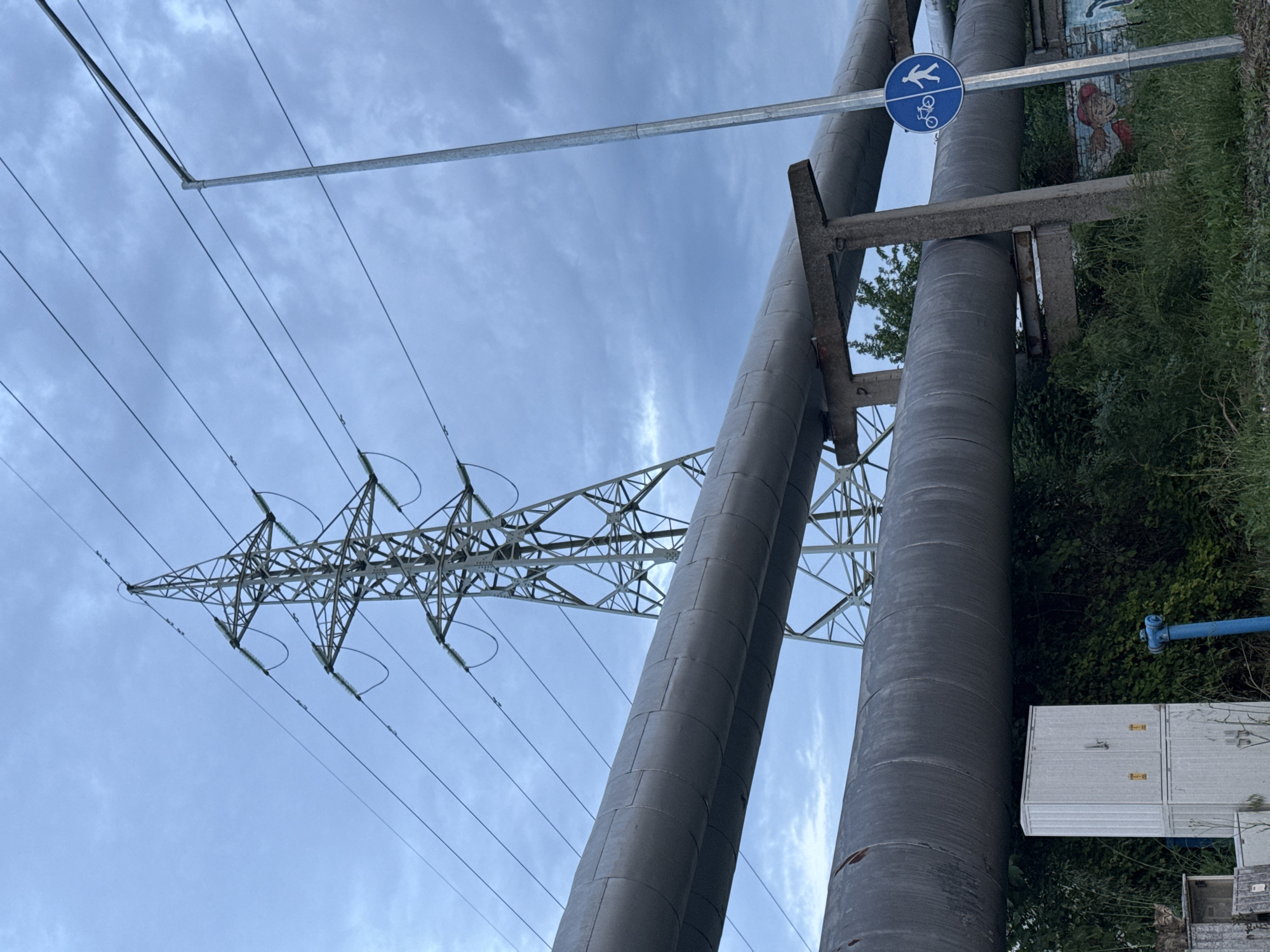 The image shows two large, gray pipes running vertically, with an electrical pylon to the left. A blue and white pedestrian/bicycle sign is affixed to a metal bar above the pipes. Behind the pipes is some greenery and part of a building with graffiti. In the lower right, there is a white electrical box. The sky is overcast. The location is Burgas, Bulgaria, identifiable by the distinctive style of pipes and related infrastructure commonly found in the area.