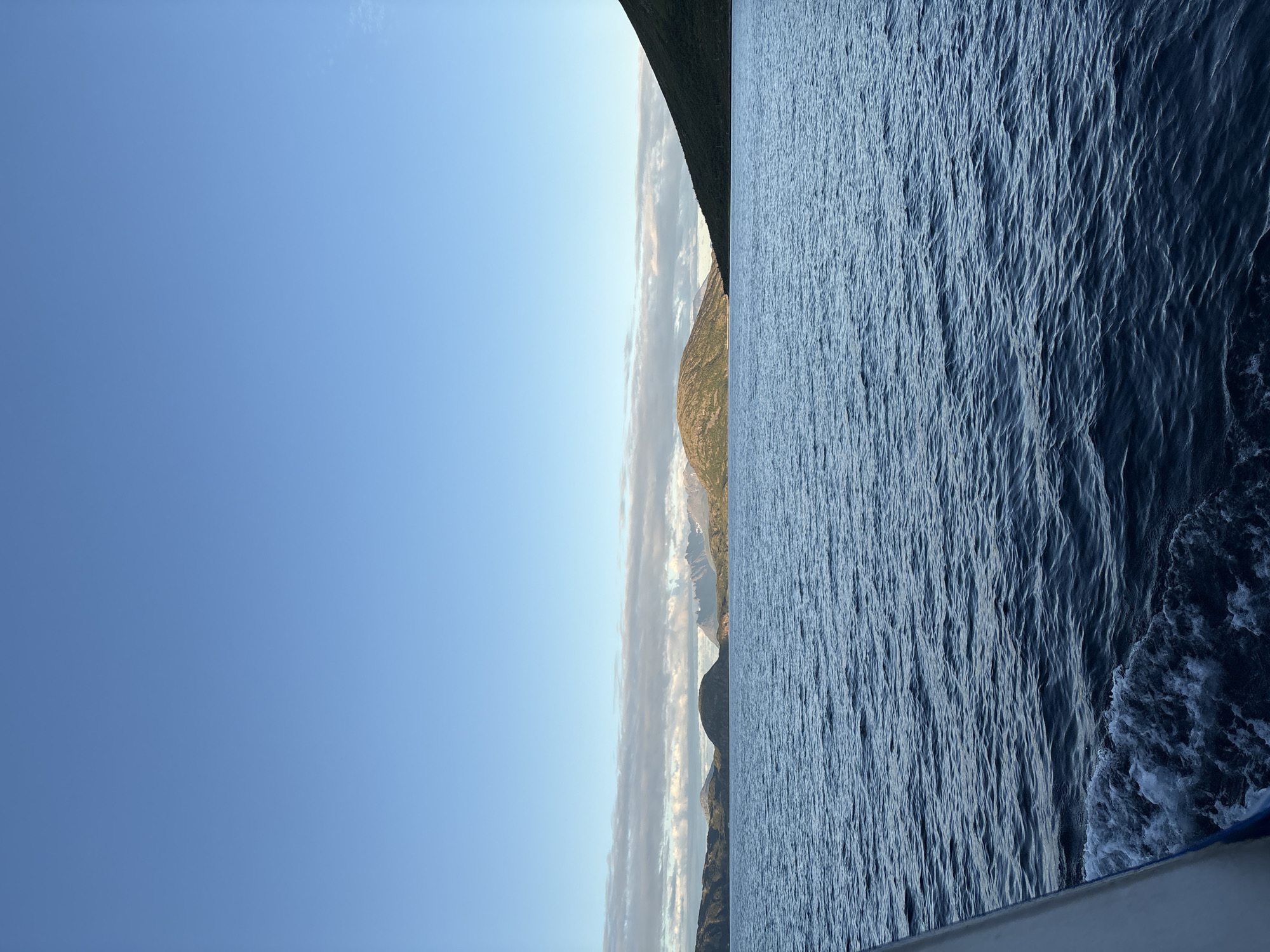 The image depicts a serene seascape with a clear blue sky above. In the foreground, dark blue water shows gentle ripples and a white wake trails from the left, indicating movement, likely from a boat. The middle ground is dominated by the expansive ocean stretching towards a distant coastline. The coastline features rolling, green-topped hills and mountains, with some areas appearing rocky and barren. A strip of thin, wispy clouds runs horizontally across the horizon, separating the sea from the sky. The light suggests it is either early morning or late afternoon, with a soft, diffused sunlight illuminating the landscape. The scene evokes a sense of tranquility and natural beauty characteristic of coastal Norway. The specific location of Arnøyhamn, Norway, can be inferred from the distinct geographical features visible, such as the rugged mountains meeting the sea.