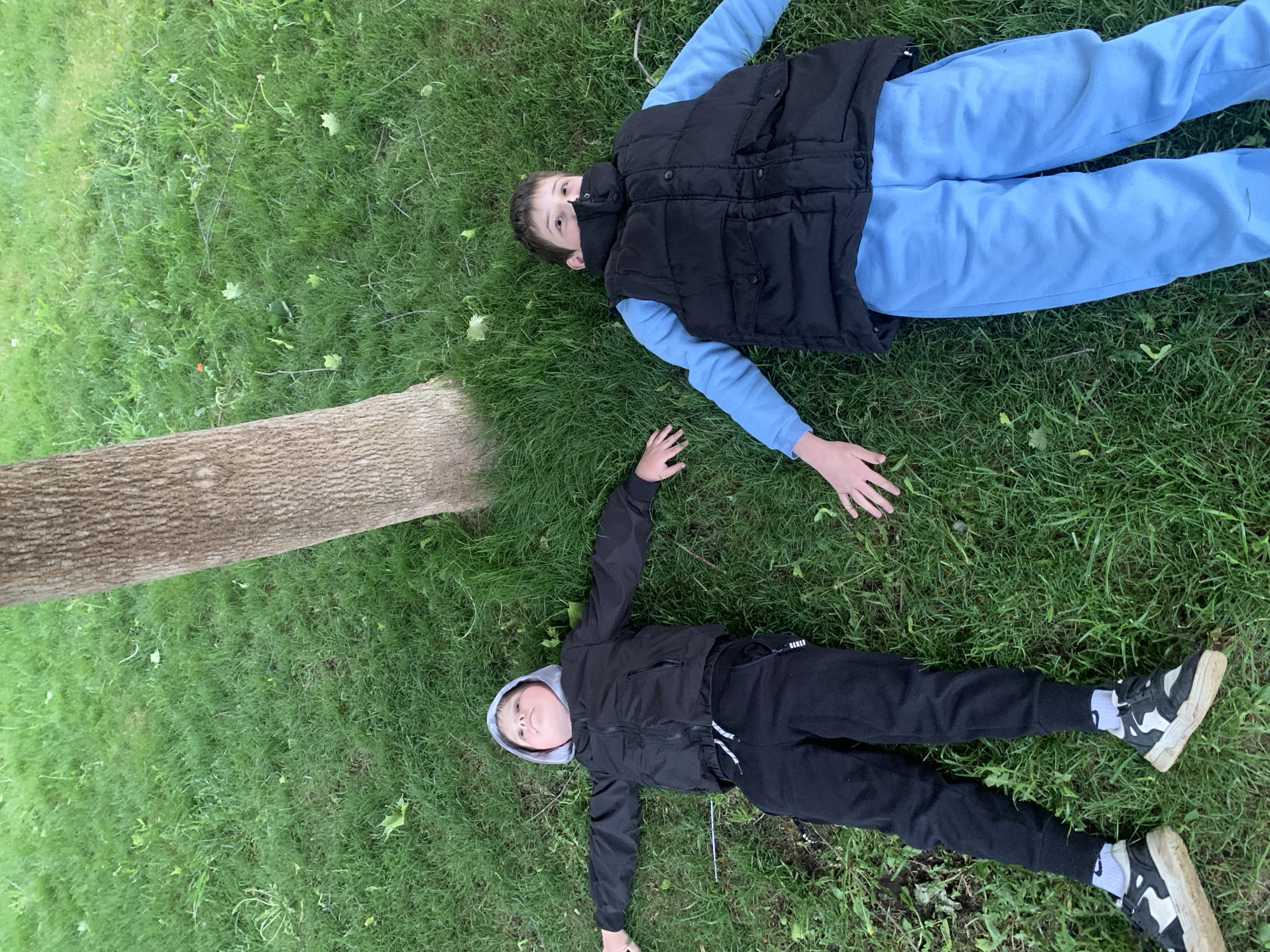 Two children lie on their backs on a grassy area. The child on the right wears a black vest over a light blue long-sleeved shirt and light blue sweatpants. The child on the left wears a black jacket and black sweatpants with white drawstrings and sneakers. A tree trunk lies horizontally between the two children. The scene appears to be outdoors during daylight hours, potentially in a park. It is not possible to ascertain the exact location as Chaikovsky, Russia from the given information.