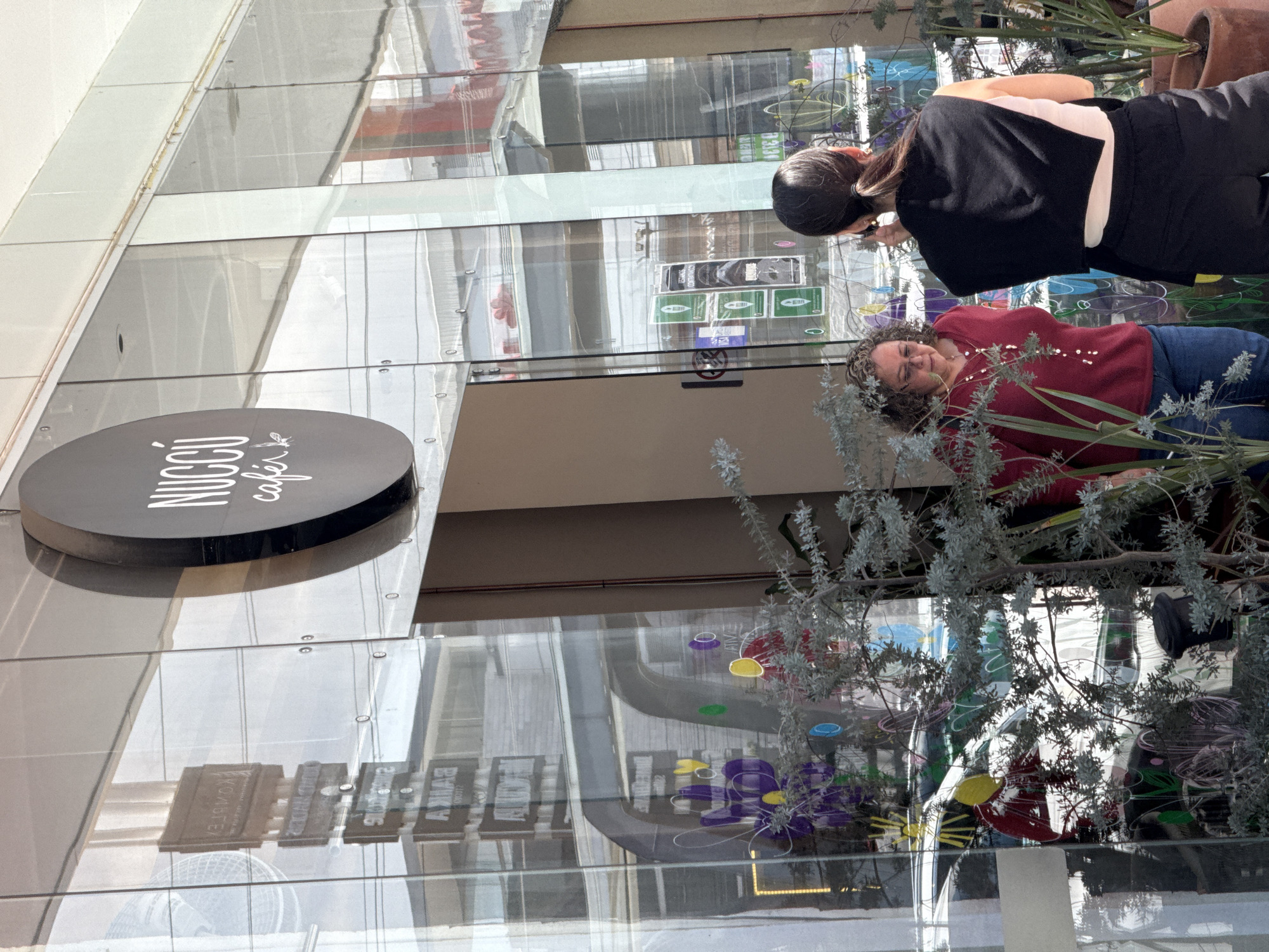 The image depicts the exterior of a cafe in Puebla, Mexico. A sign reads "NUCCI cafe". Two women stand near the entrance. One woman wears a red sweater and jeans. The second woman is in a black top and pants. Decorative plants are present. The cafe's glass facade reflects objects and light.