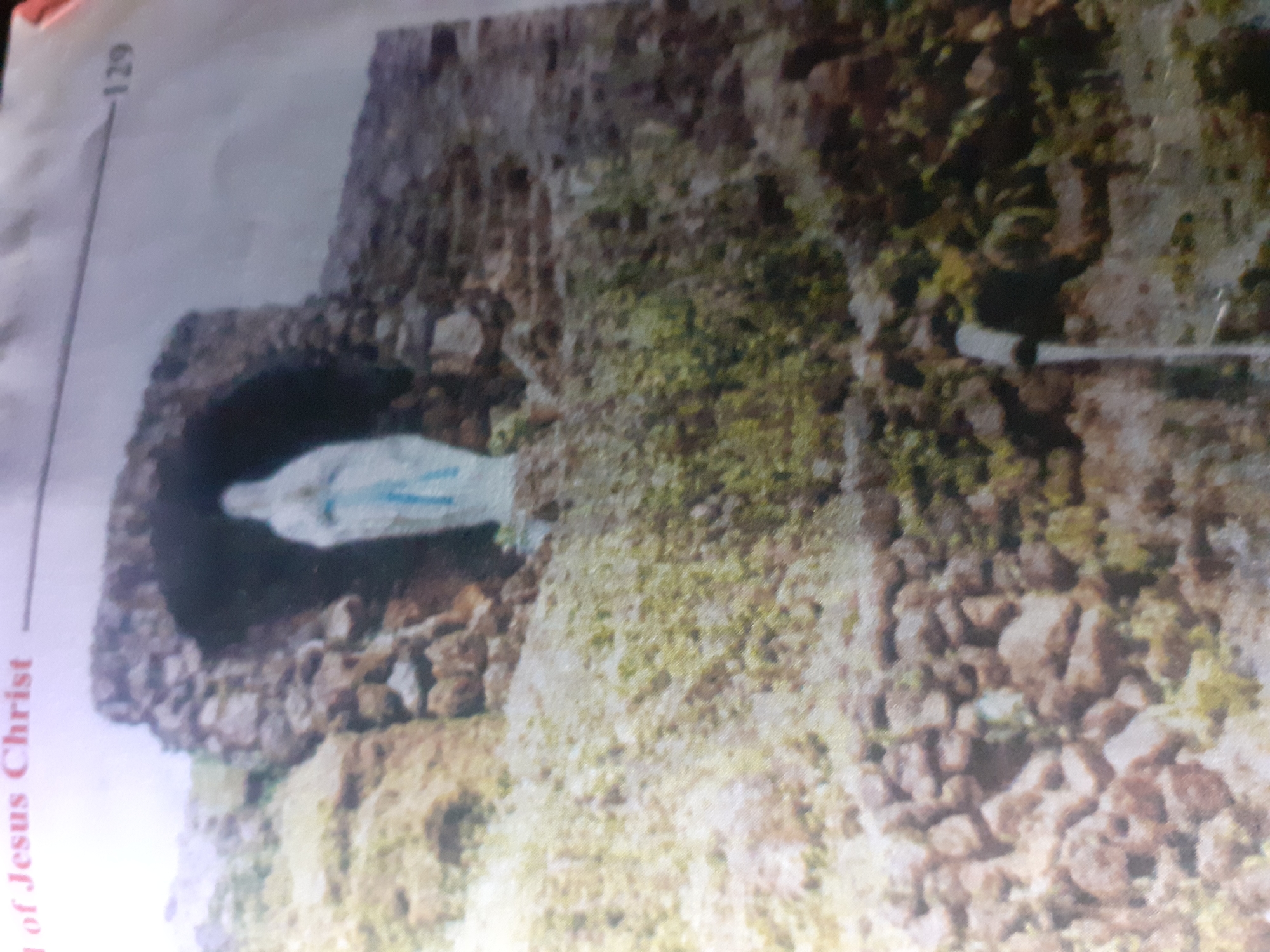 The image depicts a statue of the Virgin Mary situated within a grotto constructed of rocks. The statue is white with blue accents. A water pipe or fountain is visible in the foreground. The background consists of a rocky, overgrown landscape. Text at the top of the image reads "of Jesus Christ" and a number "129" is visible on the right side, suggesting this is a page from a book or publication. The specific location in Jalingo, Nigeria is not visually identifiable from the image itself.