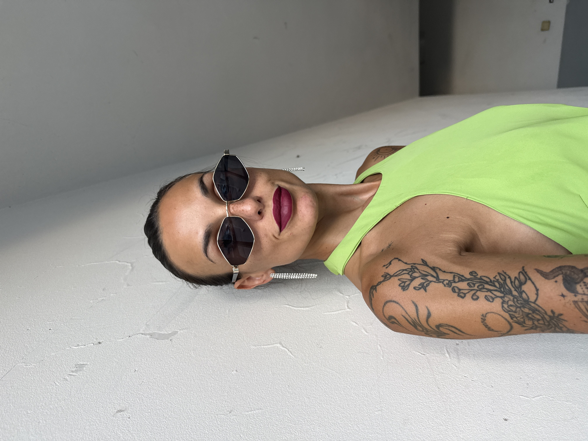 The image shows a woman with dark hair pulled back, wearing large, angular sunglasses with silver frames and a bright lime green sleeveless top. She has dramatic dark red lipstick and long, dangling crystal earrings. She is positioned in front of a textured white wall, leaning slightly to her left. Her left arm, visible on the left side of the frame, is adorned with several black tattoos, including floral and abstract designs. The background shows a dimly lit corridor or room with a grey floor. The scene appears to be indoors. The location of Pleven, Bulgaria is not evident from the image itself.