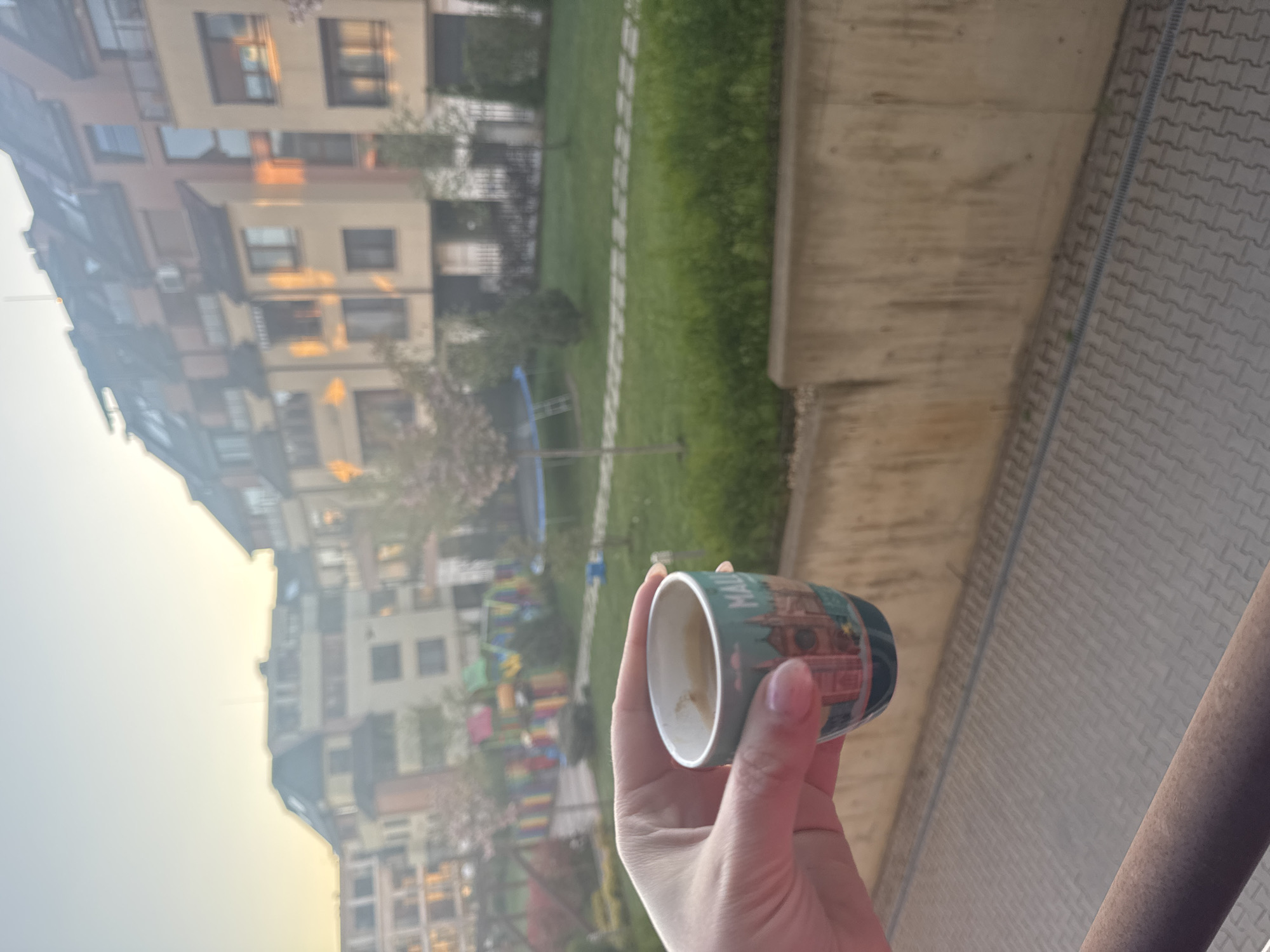 A first-person perspective image taken from a balcony in Sofia, Bulgaria, shows a hand holding a paper cup with a depiction of the Alexander Nevsky Cathedral, a landmark in Sofia. The cup contains leftover coffee. In the background, a modern residential building complex is visible across a green lawn with a playground. The balcony appears to overlook a paved area. The photo was likely taken in the early morning or late afternoon, judging by the soft lighting.