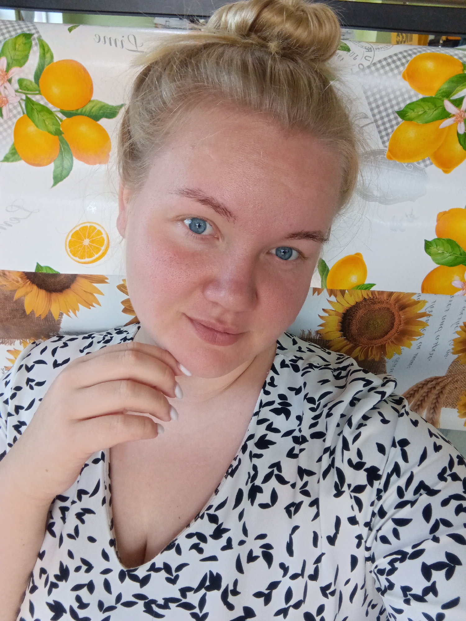 A close-up, selfie-style photograph shows a young woman with blonde hair pulled into a bun on top of her head. She has blue eyes and fair skin with some redness on her cheeks. She is wearing a white and black patterned blouse. Her left hand is resting on her chin, and her fingernails are painted white. The background of the image features a tablecloth with a pattern of lemons, sunflowers, and leaves. The location of this image is not specified in the provided context, and therefore cannot be identified as being in the city of Фоки, Russia.