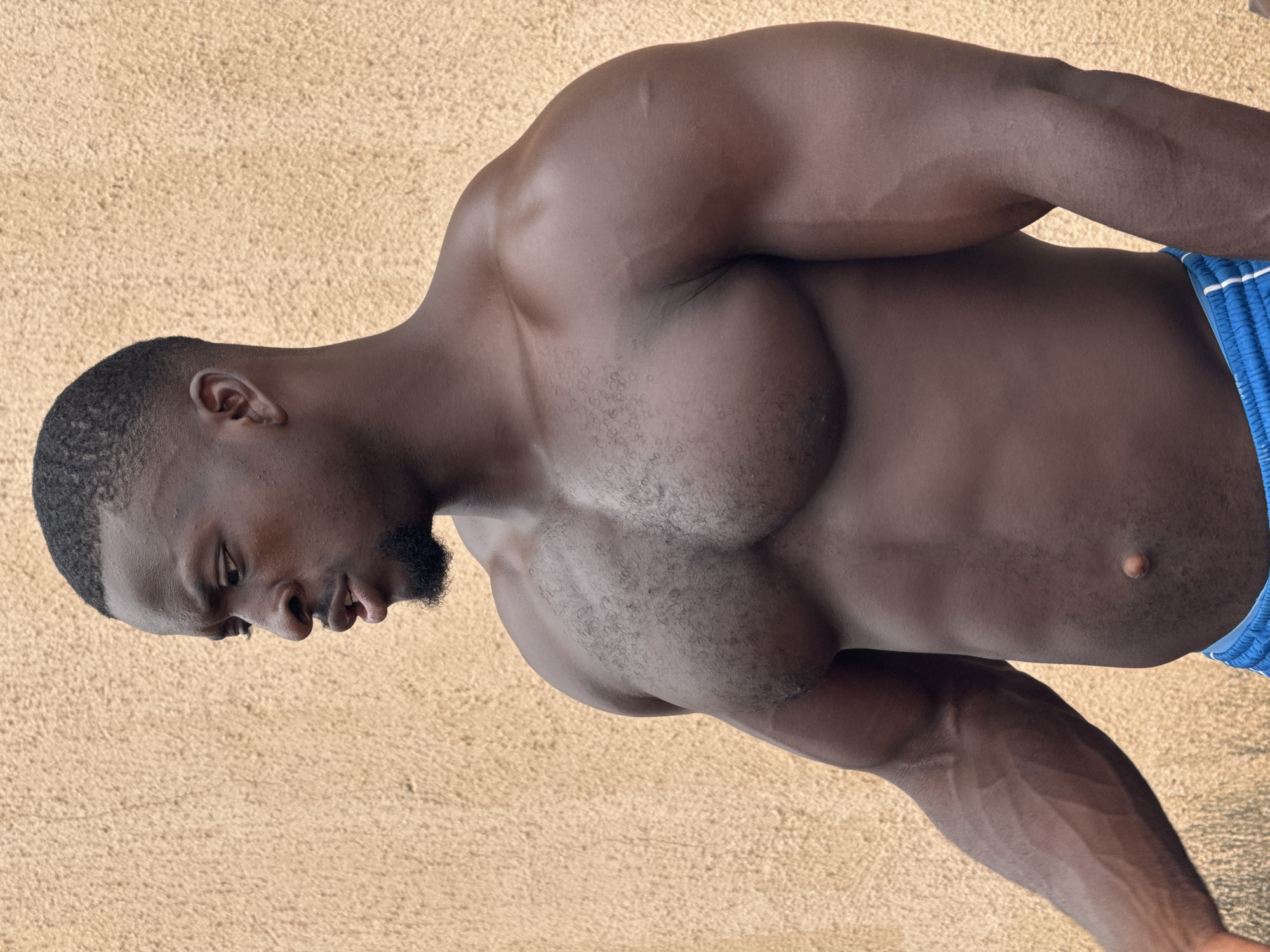 A medium shot shows a muscular Black man from the chest up, facing left and looking down and away from the camera. He has short black hair and a goatee. He is shirtless, wearing blue athletic shorts with white stripes. He stands in front of a textured, light brown wall. The lighting suggests an outdoor environment, likely during the day. The scene appears to be in Akwanga, Nigeria.