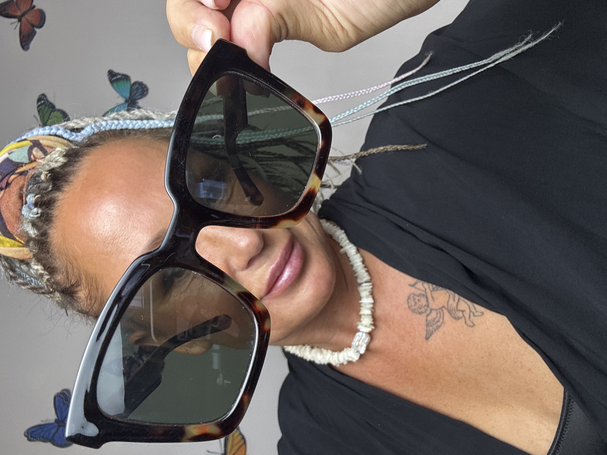 A medium shot shows a woman holding large, tortoiseshell-patterned sunglasses up to her face. She has a colorful bandana tied around her head and her hair is in braids with colorful strands. She is wearing a black garment and a white shell necklace. There is a tattoo of an angel on her chest. The background is a gray wall with several butterfly decals. The image is taken from a slightly low angle, and the woman's face is partially obscured by the sunglasses.