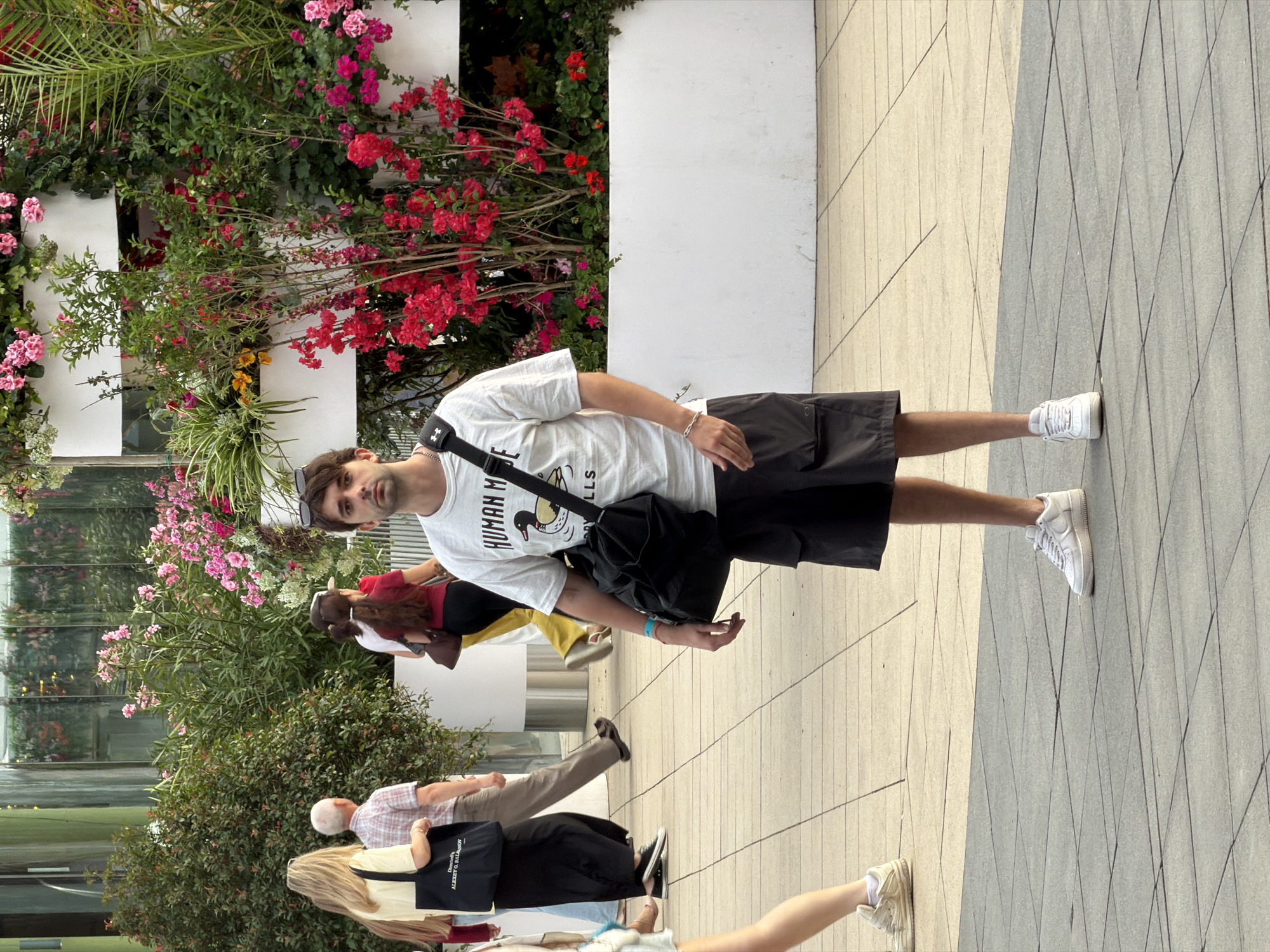 A man stands in the foreground of an outdoor scene, facing the camera. He is wearing a white t-shirt with a graphic of a duck and text, black cargo shorts, and white sneakers. A black shoulder bag is slung across his chest. He has short, dark hair and is wearing sunglasses on his head.
Behind him, several other people are walking. To the left, a woman with long blonde hair wears a yellow shirt and carries a black tote bag. Next to her, an older man in a plaid shirt walks away from the camera. Further back, a woman with dark hair wears a red top and dark pants.
The background features a vertical garden with abundant pink and red flowers, as well as lush green foliage. This garden is contained within white planters. The ground is paved with large, square tiles, with distinct grout lines creating a grid pattern. The scene appears to be in a public outdoor space, likely a pedestrian area or walkway within a city. The location is identified as Moscow, Russia.
