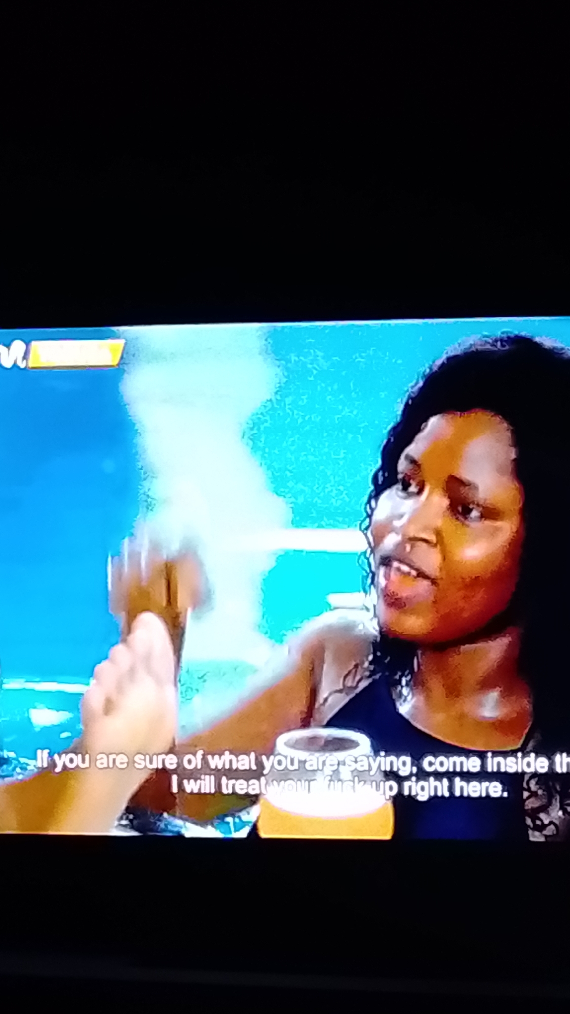 The image is a still frame from a video, displaying a woman with dark curly hair and dark skin speaking. She is looking slightly to her left and her mouth is open as if mid-sentence. She is wearing a dark top with some beading or embellishment on the strap. To her left, a hand is visible reaching out, fingers splayed. Subtitles are displayed at the bottom of the frame, partially obscured by a graphic element that appears to be a stylized depiction of a beer or drink with a frothy top. The background is blurred and appears to be an indoor setting with blue and green tones. The overall image quality is slightly grainy, suggesting it was captured from a screen. The location in Lokoja, Nigeria is not discernible from the visual information provided in the image.