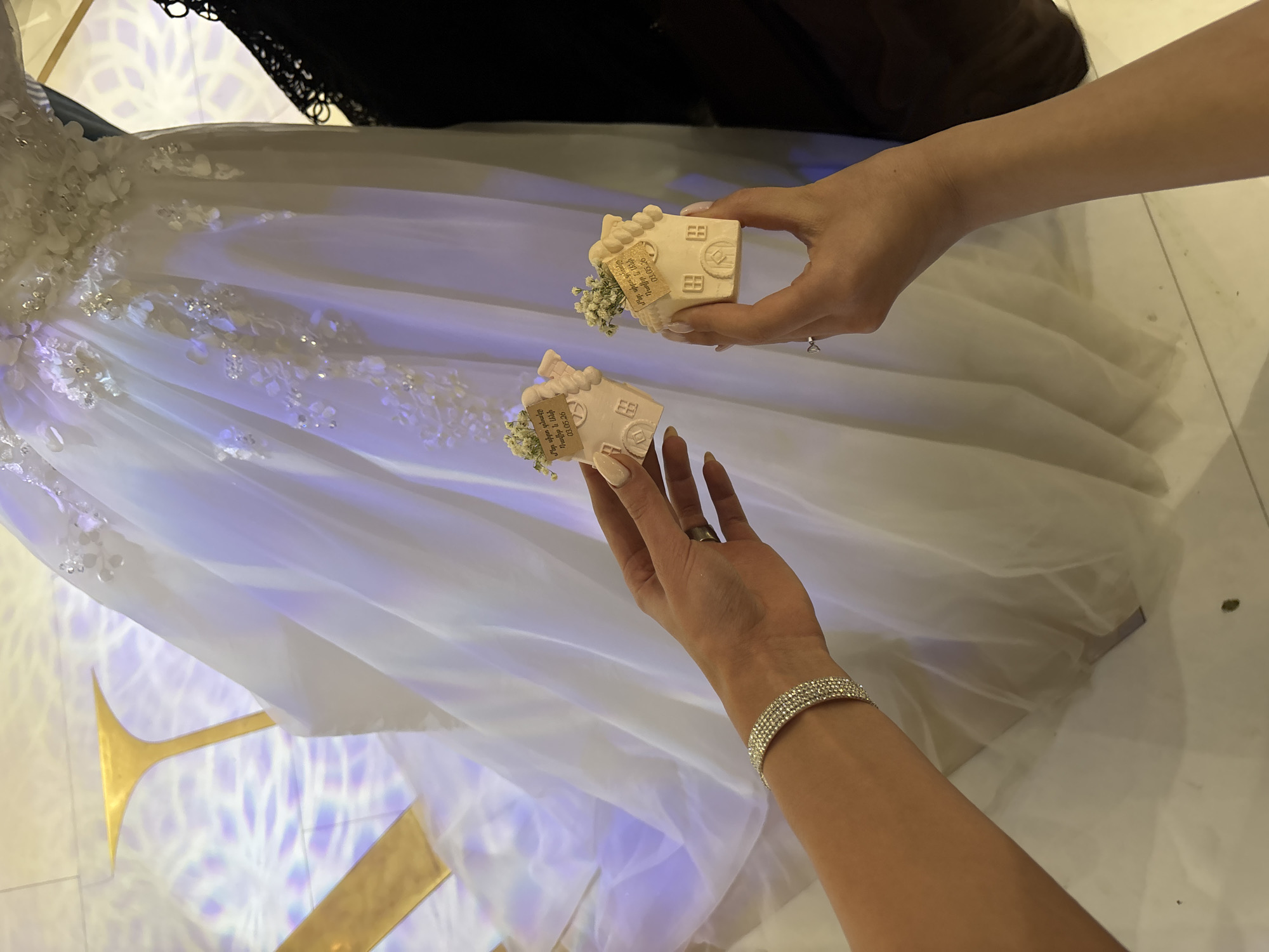 Two female hands hold small, house-shaped favors with tags attached, positioned against the backdrop of a white wedding dress. The favors are beige, and adorned with tiny white flowers and brown labels. The hands are light-skinned, with manicured nails and jewelry; one hand has a ring, and the other a bracelet. The wedding dress is detailed with floral embellishments and is draped over what appears to be a golden support structure. The scene is illuminated with a pattern of geometric shapes, cast on the floor, and suggests an indoor setting. The location of Masis, Armenia is not directly discernible from the image, but the context of a wedding, favors written in Armenian, and the reference to Masis implies a connection.