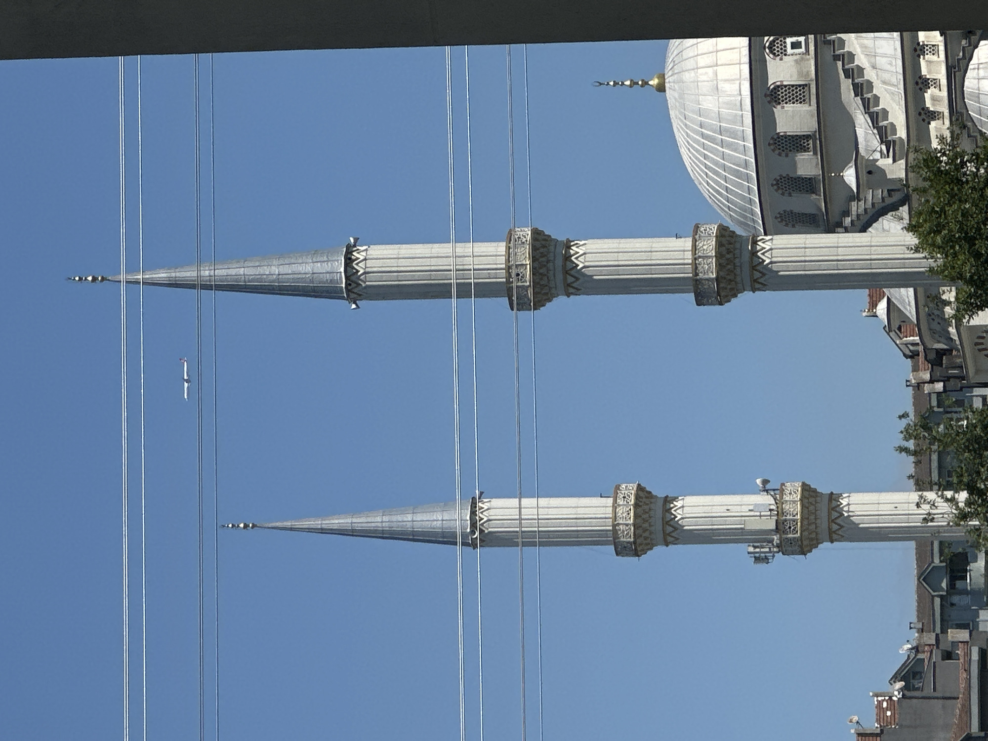 The image shows two minarets of a mosque, a dome, and an airplane in the sky. The minarets are tall and slender, with decorative bands around their bases and tops. The dome is large and white, with arched windows. The airplane is small and white, and appears to be flying at a high altitude. The scene is set against a clear blue sky. In the foreground, there are power lines stretching across the frame. The mosque is located in Esenyurt, Turkey. The buildings in the background suggest an urban environment.