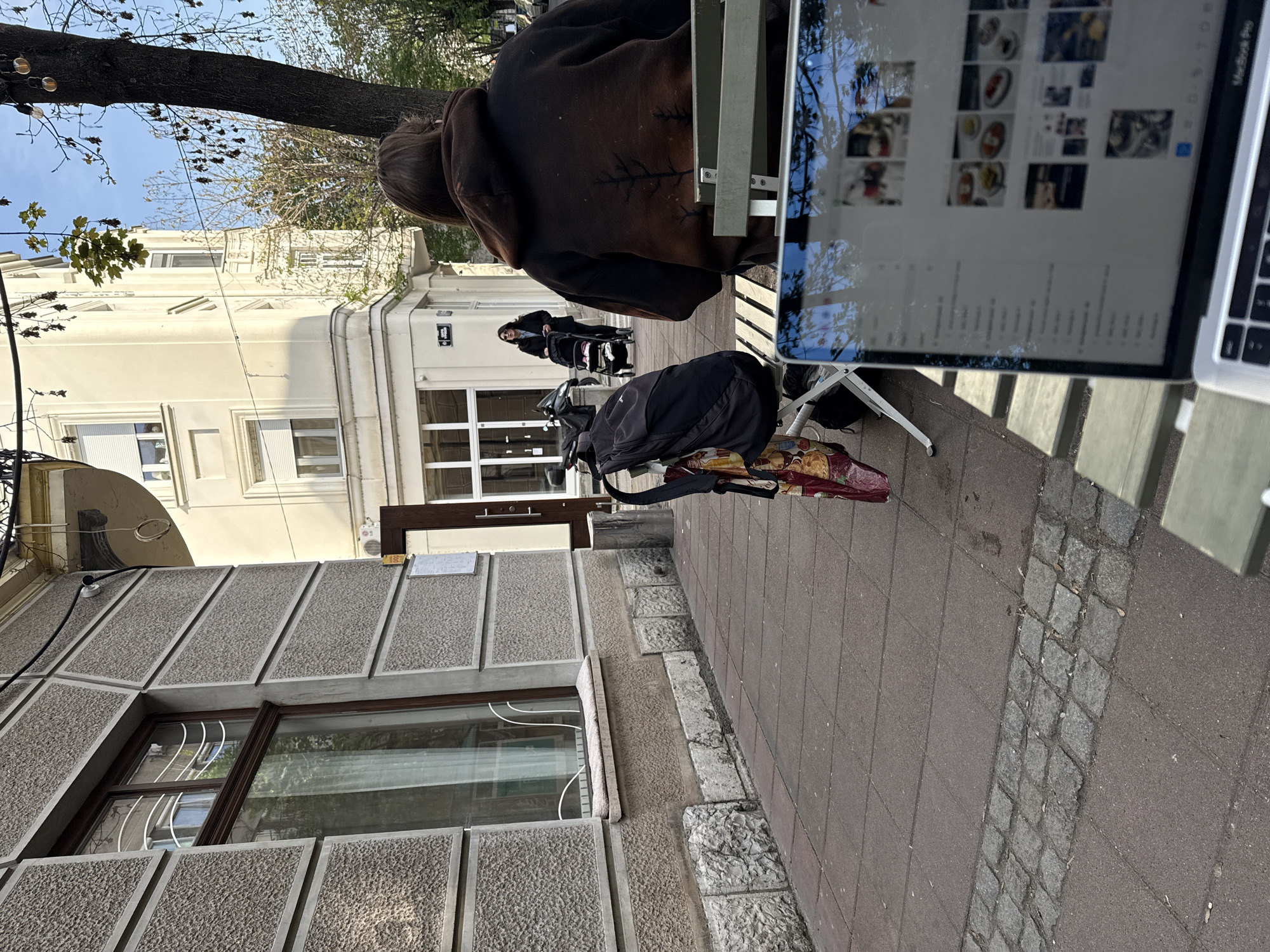 An outdoor urban scene is captured at an angle, featuring a person wearing a dark brown hooded jacket seated at a metallic folding table, with a laptop open on the table, the screen displaying a grid of images. Adjacent to the table, a black backpack rests on the ground. In the background, a woman pushes a stroller along the sidewalk.
The setting appears to be on a street in Belgrade, Serbia. Architectural details are evident on the left, including a building with gray textured walls and a window featuring white bars. In the background, a cream-colored building is partially visible. The street is paved with red tiles, interspersed with a strip of gray cobblestones.