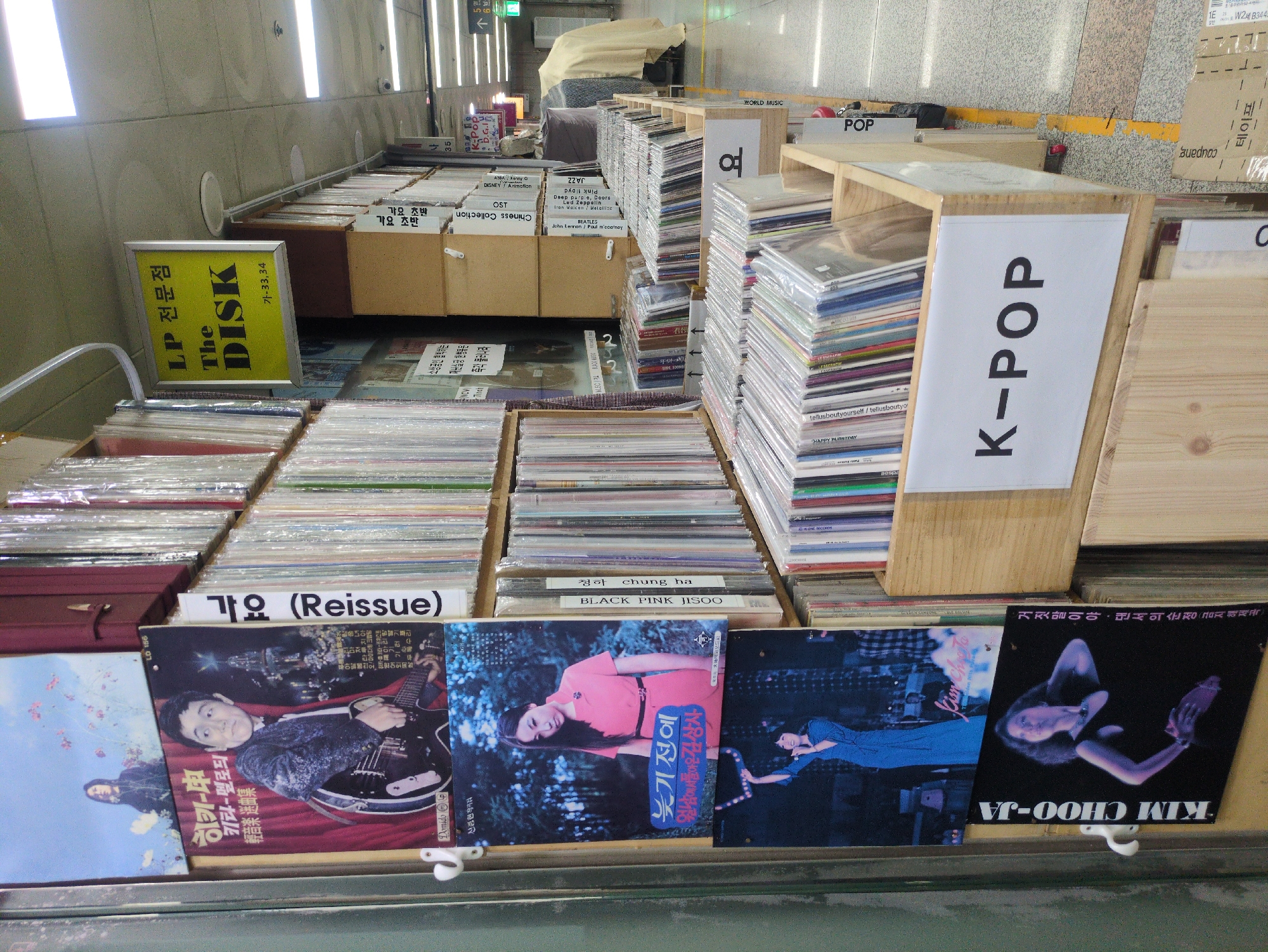 This image captures a detailed view of an LP record store, "The DISK," located in an underground shopping area in Seoul, South Korea. The store is identified by a yellow hanging sign reading "LP 전문점 The DISK" (LP specialty store The DISK) with stall numbers "가-33,34."
The scene is dominated by shelves and wooden crates filled with numerous vinyl records, many encased in clear plastic sleeves. On the left, several oversized album covers are displayed vertically. The topmost visible cover features two figures in a field of flowers. Below it, a male musician is depicted holding a guitar against a dark background, with Korean text "히키-쏭" (Hiki-ssong) and "기타-멜로디" (Guitar-Melody) visible. Further down, a female artist in a pink dress stands in a forest, with the album title "늦기 전에" (Before It's Too Late). The bottommost visible cover shows a female artist identified as "KIM CHOO-JA" (김추자) in a black dress against a black background.
Records are organized into genres, indicated by white labels on the wooden crates and shelves. Visible genre labels include "K-POP," "가요 (Reissue)" (Korean pop/folk music - Reissue), "Chinese Collection," "OST," "Disney/Animation," "BEATLES," "Pink Floyd," "JAZZ," and "WORLD MUSIC." Specific album spines mentioning "BLACK PINK JISOO" and "청하" (Chungha) are also discernible.
The environment is an indoor, brightly lit commercial space. The floor is tiled with a gray and white pattern, and a yellow demarcation line runs along the aisle. The ceiling features recessed fluorescent lighting fixtures and visible security cameras (CCTV). In the background, stacked cardboard boxes, some labeled "coupang" and "테이프" (tape), and a draped object are visible. A distant sign in Korean indicates "5번 출구" (Exit 5), confirming the location within a subway station or an underground passage connected to one. No individuals are directly visible engaging with the records, but the store appears operational.