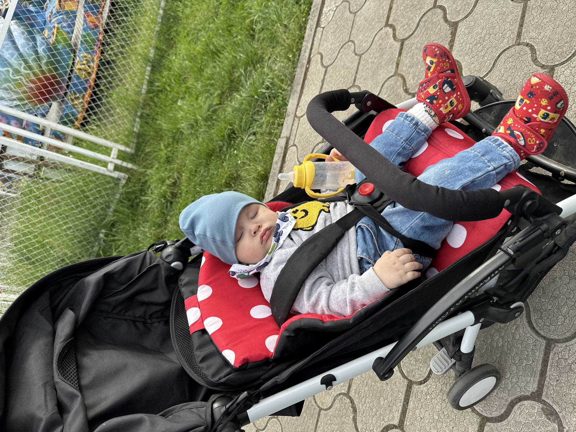 An overhead-angle medium shot depicts a sleeping baby situated within a black stroller on a paved pathway. The baby, wearing a light blue beanie, a grey long-sleeved shirt with a yellow smiling patch, a white patterned bib, blue jeans, and red shoes adorned with cartoon vehicle motifs, lies reclined in the stroller's red polka-dotted seat liner. A yellow sippy cup containing a clear liquid is held in the baby's right hand. The stroller features a black canopy, a padded black bumper bar, black harness straps, and a white/light grey frame, with one front wheel visible. The ground consists of interlocking grey hexagonal pavers. In the midground, a strip of vibrant green grass is visible. The background features a white chain-link fence, behind which colorful structures, possibly amusement park rides, are partially visible through the mesh. The scene is illuminated by natural daylight.