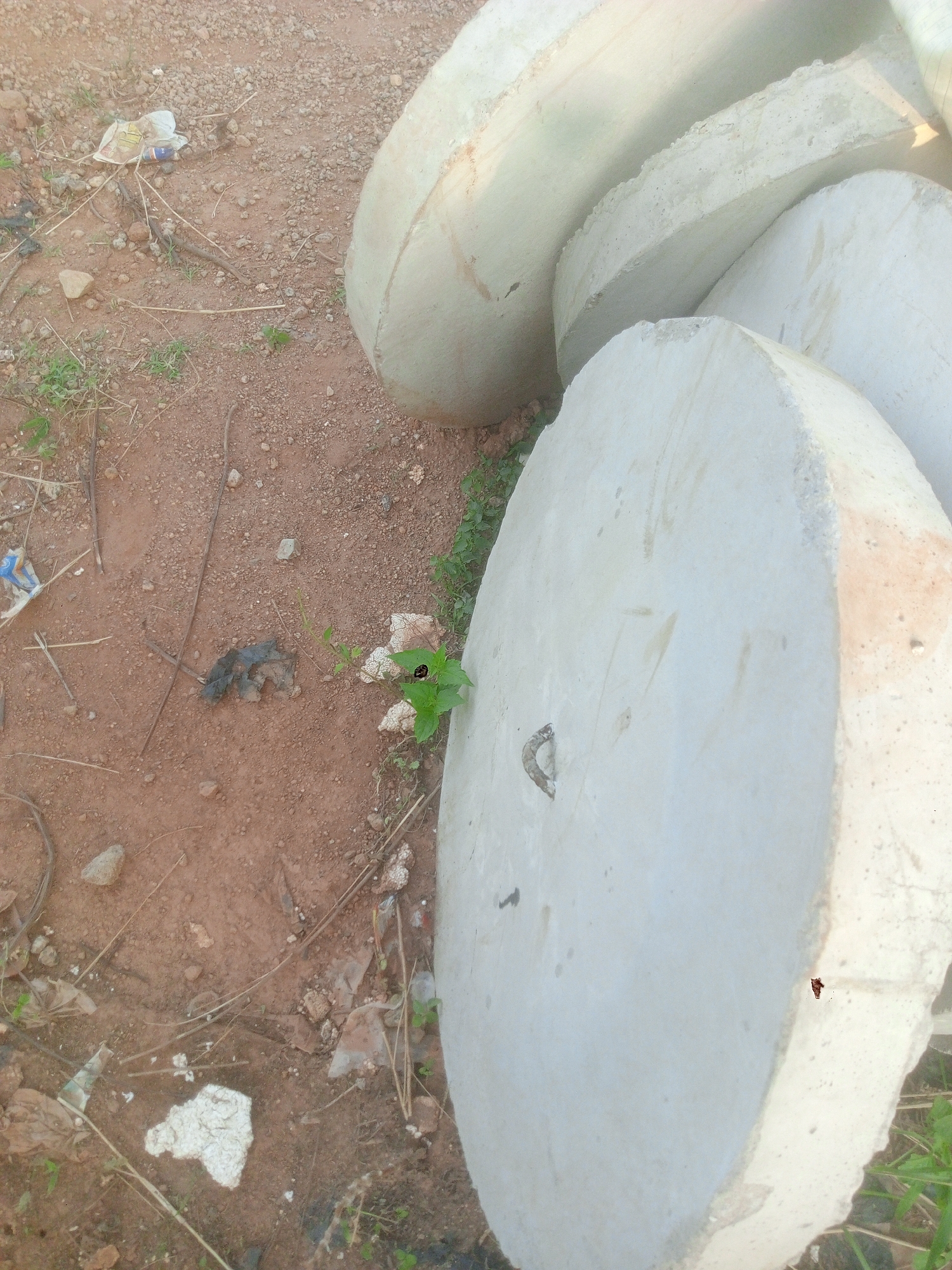 The image captures an outdoor scene dominated by several large, circular concrete slabs resting on a reddish-brown dirt ground. These heavy, light-grey slabs are stacked or leaning against each other, primarily occupying the right side and foreground of the frame. One slab in the foreground features a small, metallic, U-shaped hook or handle embedded in its surface.
The ground is composed of coarse, reddish-brown soil mixed with small stones and patches of sparse green vegetation, mostly weeds and grass. Scattered across the dirt are various pieces of debris, including dry twigs, small rocks, and discarded plastic items. A crumpled white and blue plastic wrapper is visible in the upper left corner of the dirt area, with indistinct, faint text on it. Other small fragments of white and dark plastic or paper are also interspersed on the ground.
The lighting suggests it is daytime, with bright natural light illuminating the scene, casting no distinct long shadows. The overall impression is an unkempt or industrial outdoor area, possibly a storage site for construction materials in Gwarinpa, Nigeria. There are no people visible, and no specific activity is taking place; the scene is static.