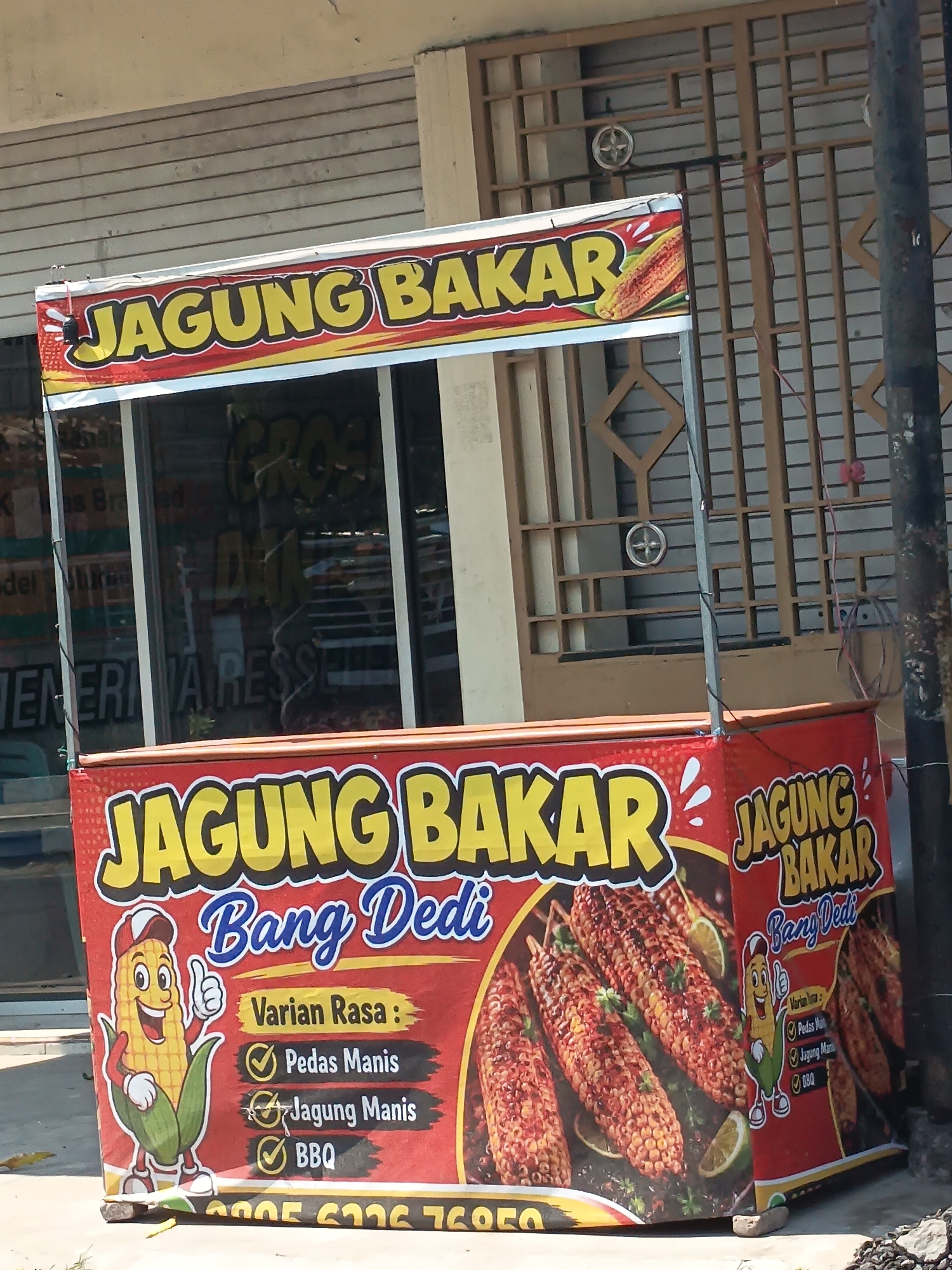 The image shows a street food stall in Kandangjati Kulon, Indonesia, selling grilled corn. The stall is decorated with a red banner featuring yellow text that reads "JAGUNG BAKAR BANG DEDI," which translates to "Grilled Corn Dedi." A cartoon character of a corn cob is displayed, giving a thumbs-up. The banner also lists flavor options: "Pedas Manis" (Spicy Sweet), "Jagung Manis" (Sweet Corn), and "BBQ." Several ears of grilled corn with spices and lime slices are depicted on the banner, along with the text "Varian Rasa" (Flavor Variants). The stall is positioned in front of a building with barred windows, suggesting an urban or semi-urban environment. The lighting indicates it is daytime, with sunlight casting shadows.