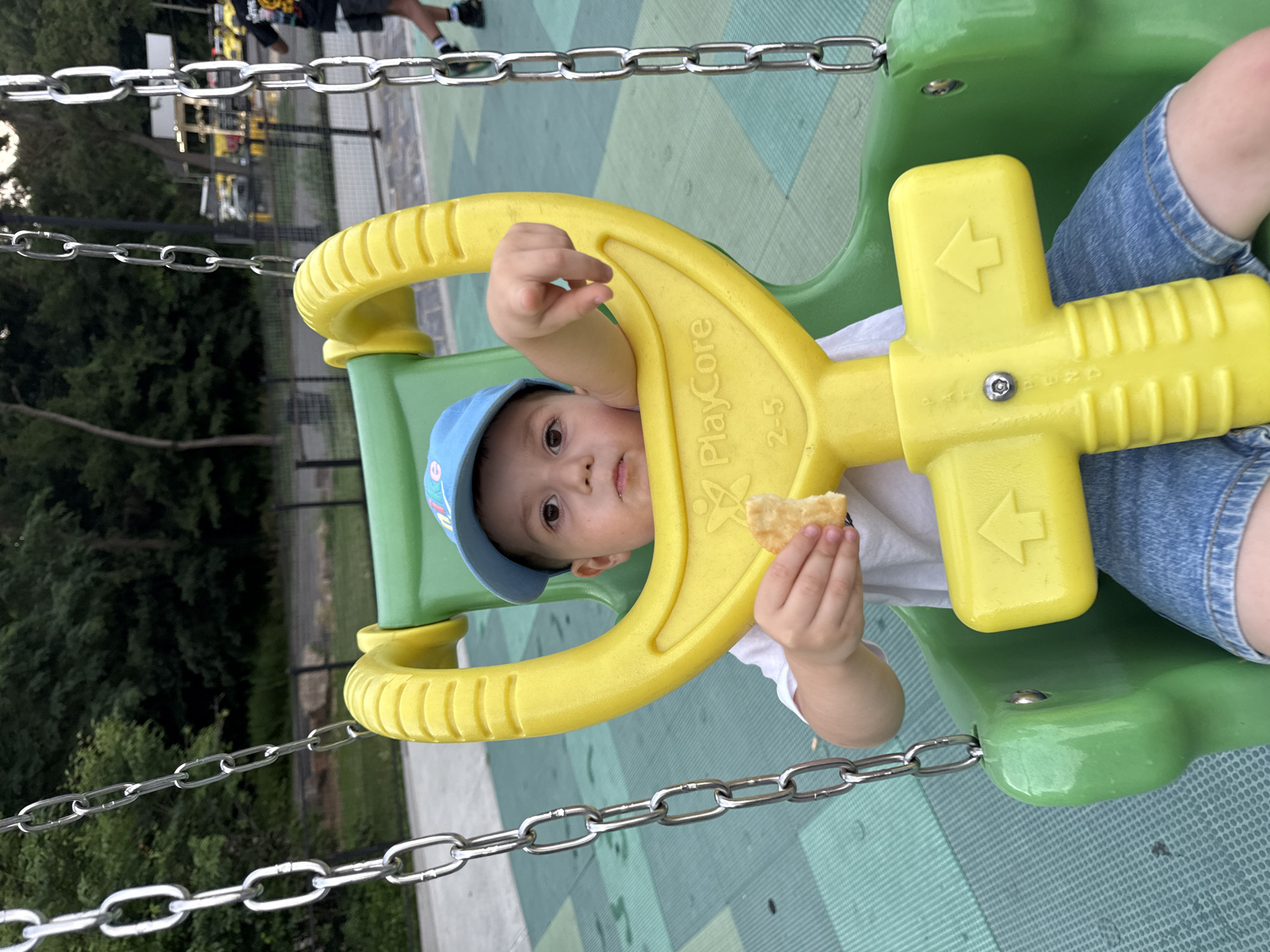 A young child wearing a blue baseball cap and a white t-shirt sits in a green and yellow swing at a playground. The child is holding a piece of food in their right hand and looking at the camera. The playground surface is a green and grey rubber mat with a patterned design. In the background, trees and a fence can be seen, as well as a road with yellow vehicles. The image appears to have been taken in New York City, United States of America.