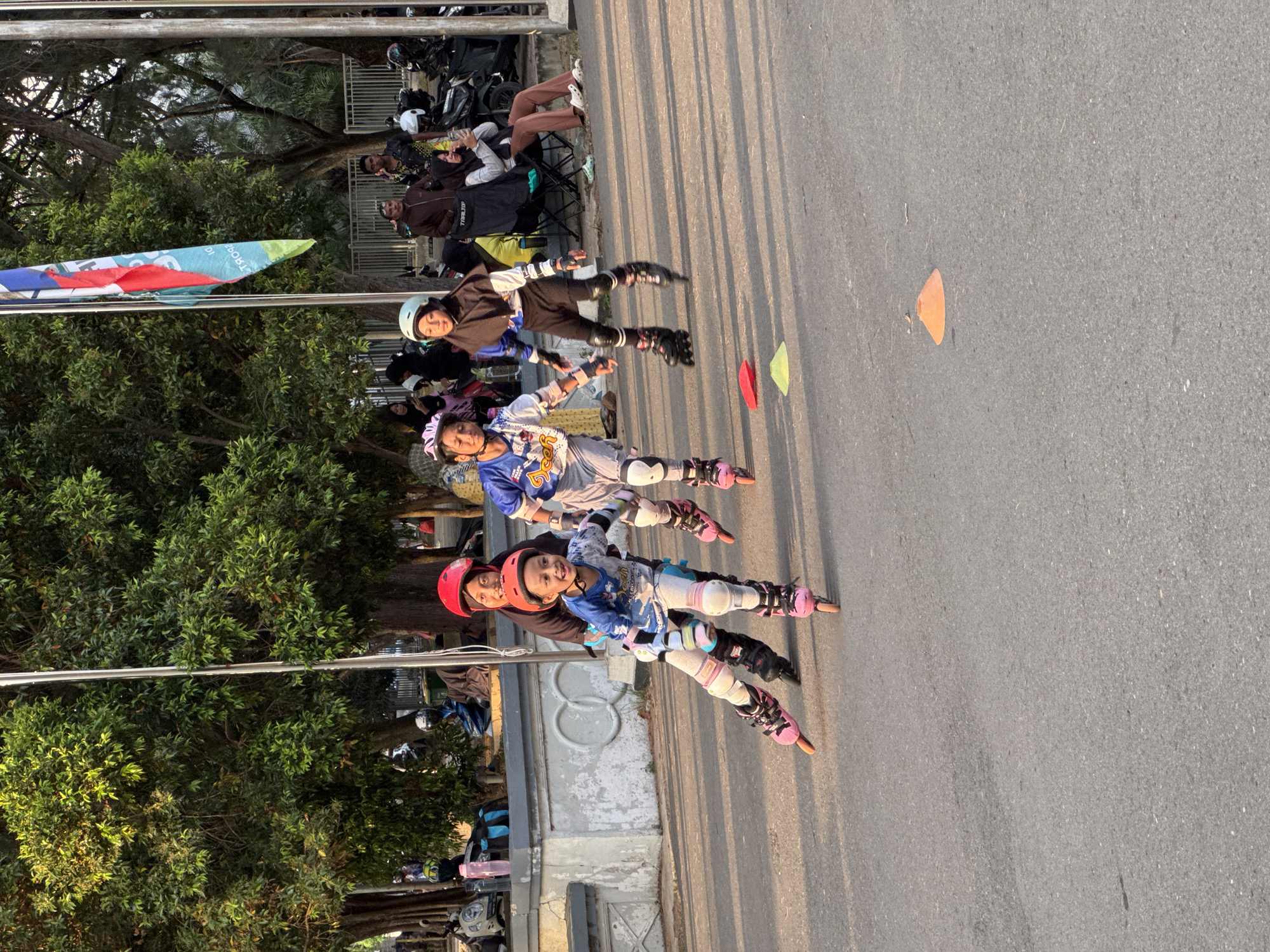 In Banda Aceh, Indonesia, on a paved outdoor area bathed in sunlight, a group of children are engaged in rollerblading. Four children are in the foreground, wearing protective gear including helmets and knee pads, and are actively skating. One child, in the front left, wears a pink helmet and blue and white rollerblades, with their mother, also wearing a red helmet, assisting from behind. Another child, to their right, sports a light blue helmet and blue and white rollerblades, appearing to be guided by an adult also wearing a helmet. Further back and to the right, two more children are rollerblading. Red and yellow traffic cones are scattered on the pavement, indicating a possible training or practice area. In the background, trees provide a lush green canopy, and a fence separates the skating area from a section with parked motorcycles and several individuals, some seated on chairs. A flagpole with a colorful flag is also visible. The overall atmosphere suggests a recreational activity taking place in a park or open public space within the city.