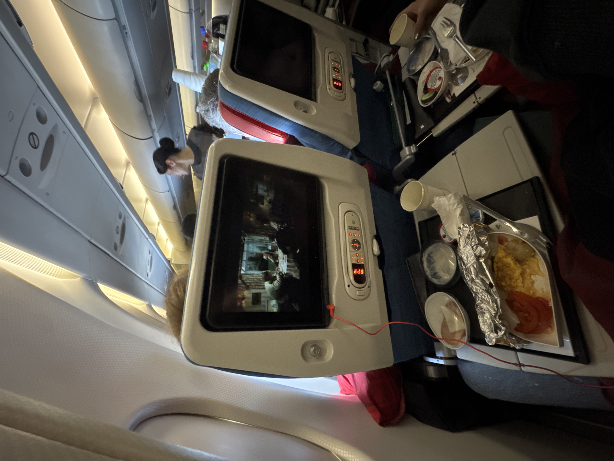 The image shows an interior view from an airplane. In the foreground, there's an in-flight entertainment screen displaying a movie, and a tray table with airline meal components, including what appears to be eggs, tomatoes, and small containers. A woman with her hair in a bun is partially visible in the background, possibly a flight attendant or another passenger. The angle indicates the photo was taken while tilting the camera. Location information points to Tambon Sa Khu, Thailand, which suggests that is the destination or departure location of the flight.