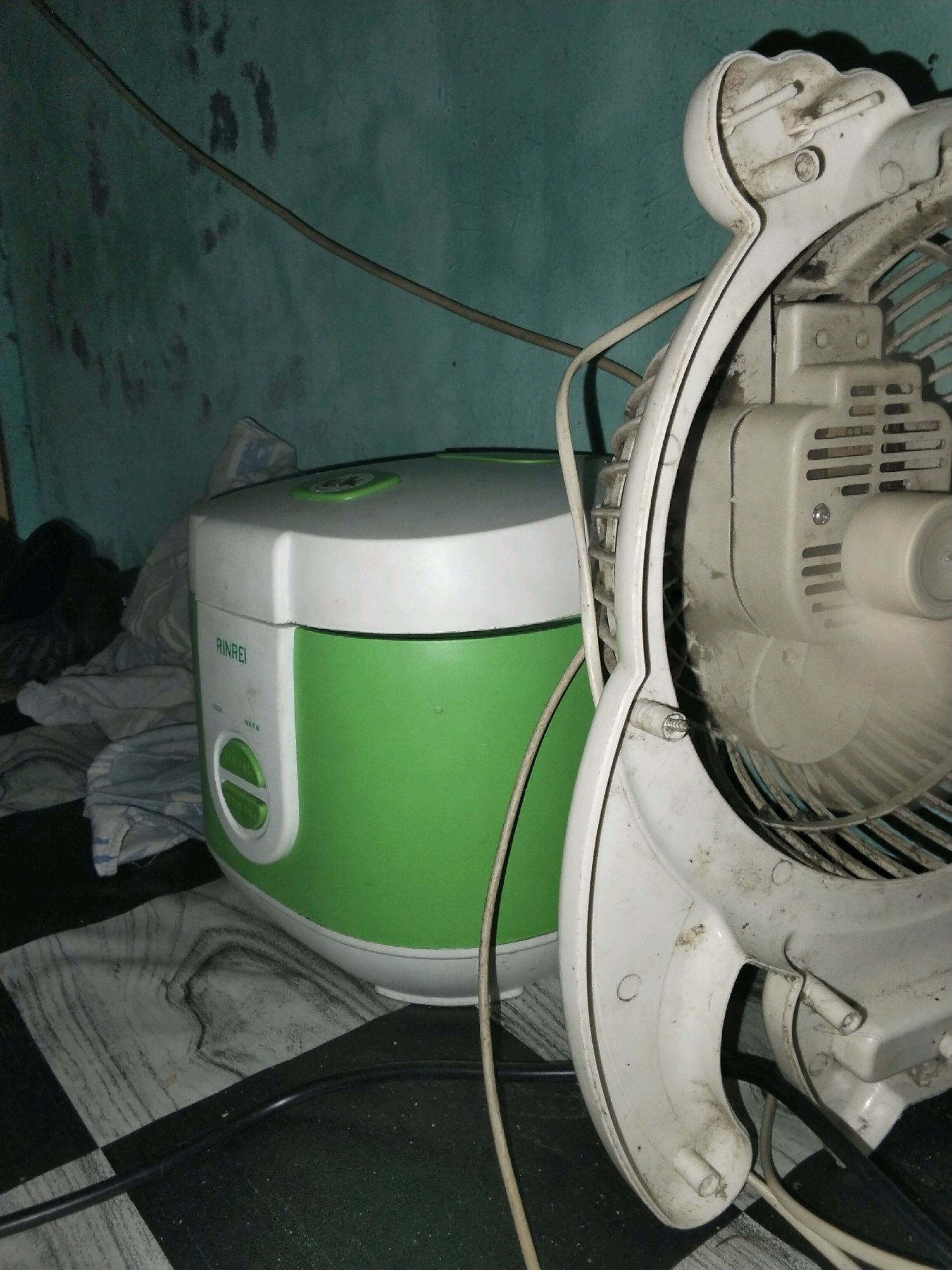 An indoor scene, likely a room in a residence in Moro, Indonesia, contains a white and green rice cooker and a dusty white electric fan. The rice cooker, positioned in the mid-left of the frame, has the brand name "RINREI" visible on its front, along with green buttons for "COOK" and "WARM". To the right of the rice cooker, the fan's casing and blades are partially visible, showing signs of dust accumulation. The background consists of a teal-colored wall with some dark marks and a power cord running horizontally. The floor in the foreground features a black and white checkered pattern with a wood-grain-like design. Several electrical cords are present, connected to the fan and possibly the rice cooker. The overall lighting is dim, suggesting it may be nighttime or the room is not well-lit. No individuals are present in the image.