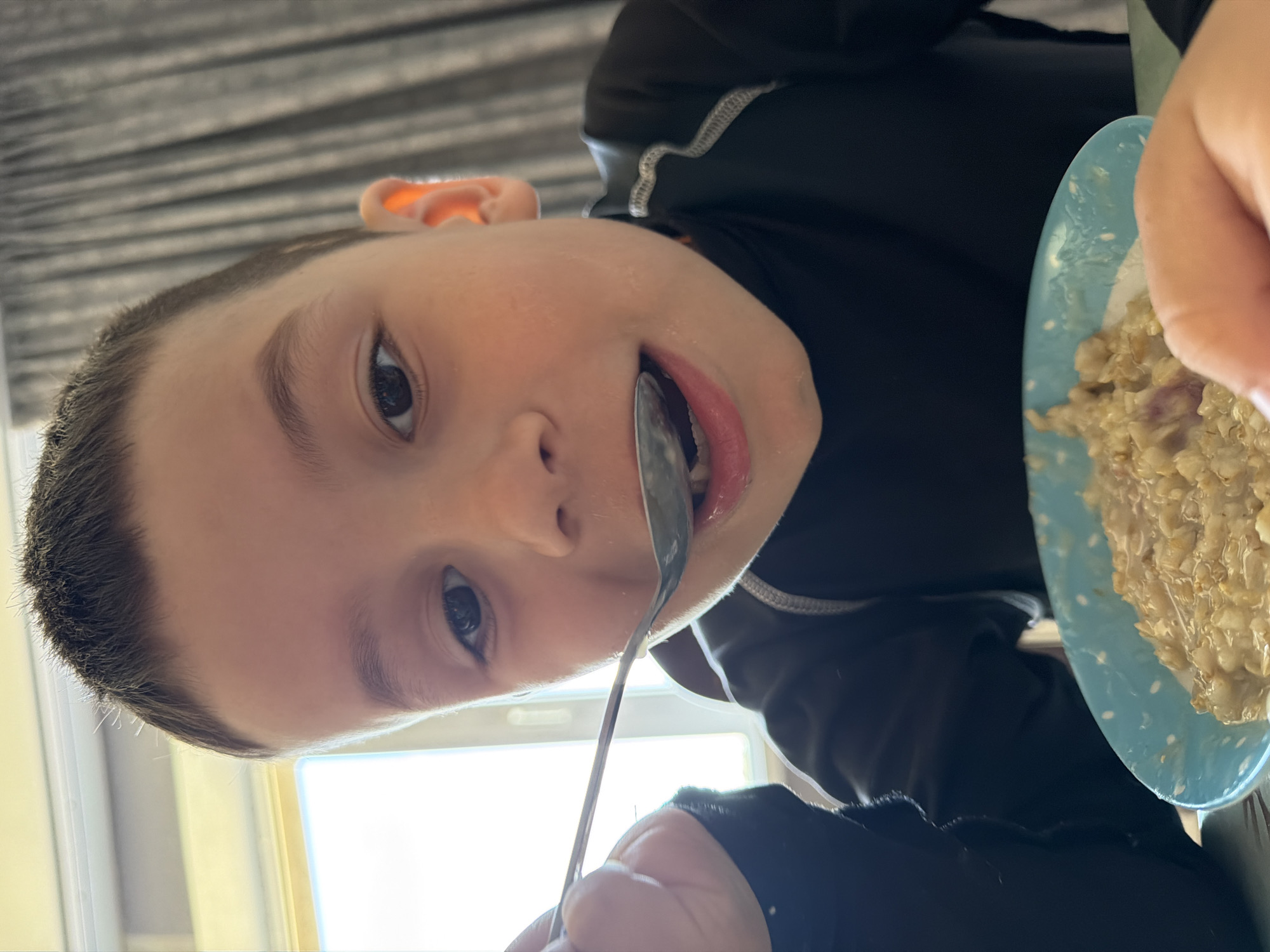 A young boy is eating oatmeal with a spoon from a blue plate. He is wearing a black long-sleeved shirt with a grey neckline. A window is visible in the background, and a window blind is present above the subject. The location is not discernible from the image, nor is there any indication it is Chaikovsky, Russia.