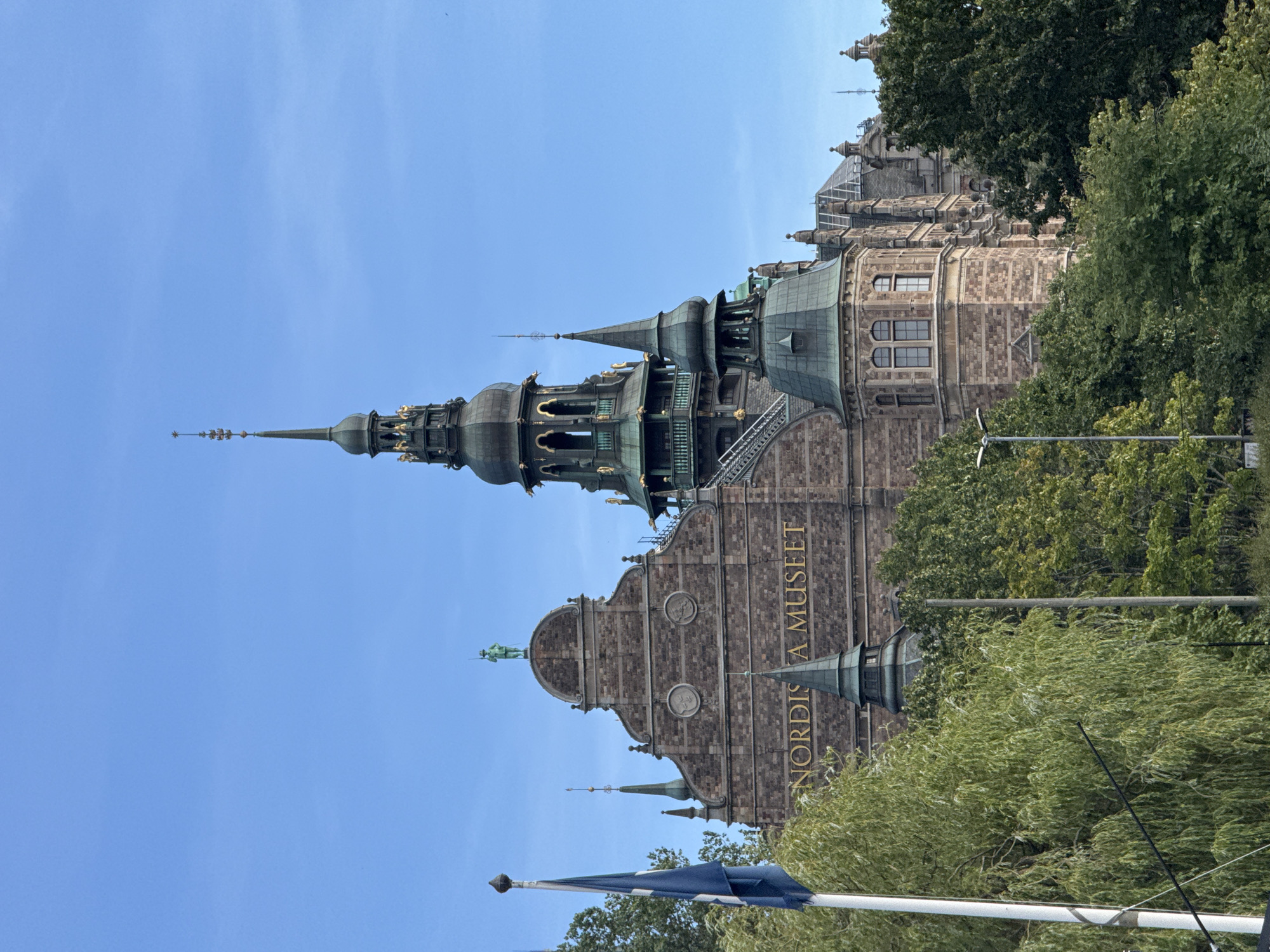 The image shows the Nordiska Museet, a museum in Stockholm, Sweden, on a clear day. The museum is a large, ornate building with stone facades and several green-roofed towers and spires. Text reading "NORDISKA MUSEET" is visible in gold lettering on the front of the building. A blue flag is flying on a flagpole to the left of the museum, partially obscured by trees. Lush green trees surround the base of the museum, with some branches extending upwards towards the building. The sky is a clear, bright blue.