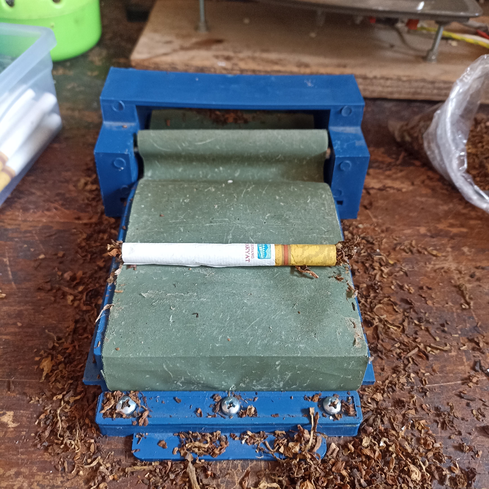 In Magelang, Indonesia, a blue cigarette rolling machine sits on a wooden surface littered with loose tobacco leaves. A single cigarette, with a white paper wrapper and a yellow filter, is positioned horizontally across the green rolling surface of the machine. The machine appears to be used, with some dust and tobacco residue visible on its blue plastic frame and green fabric roller. In the background, a plastic container holding more cigarette filters is partially visible to the left, and a clear plastic bag filled with tobacco is to the right. The overall scene suggests a setting for manual cigarette production.