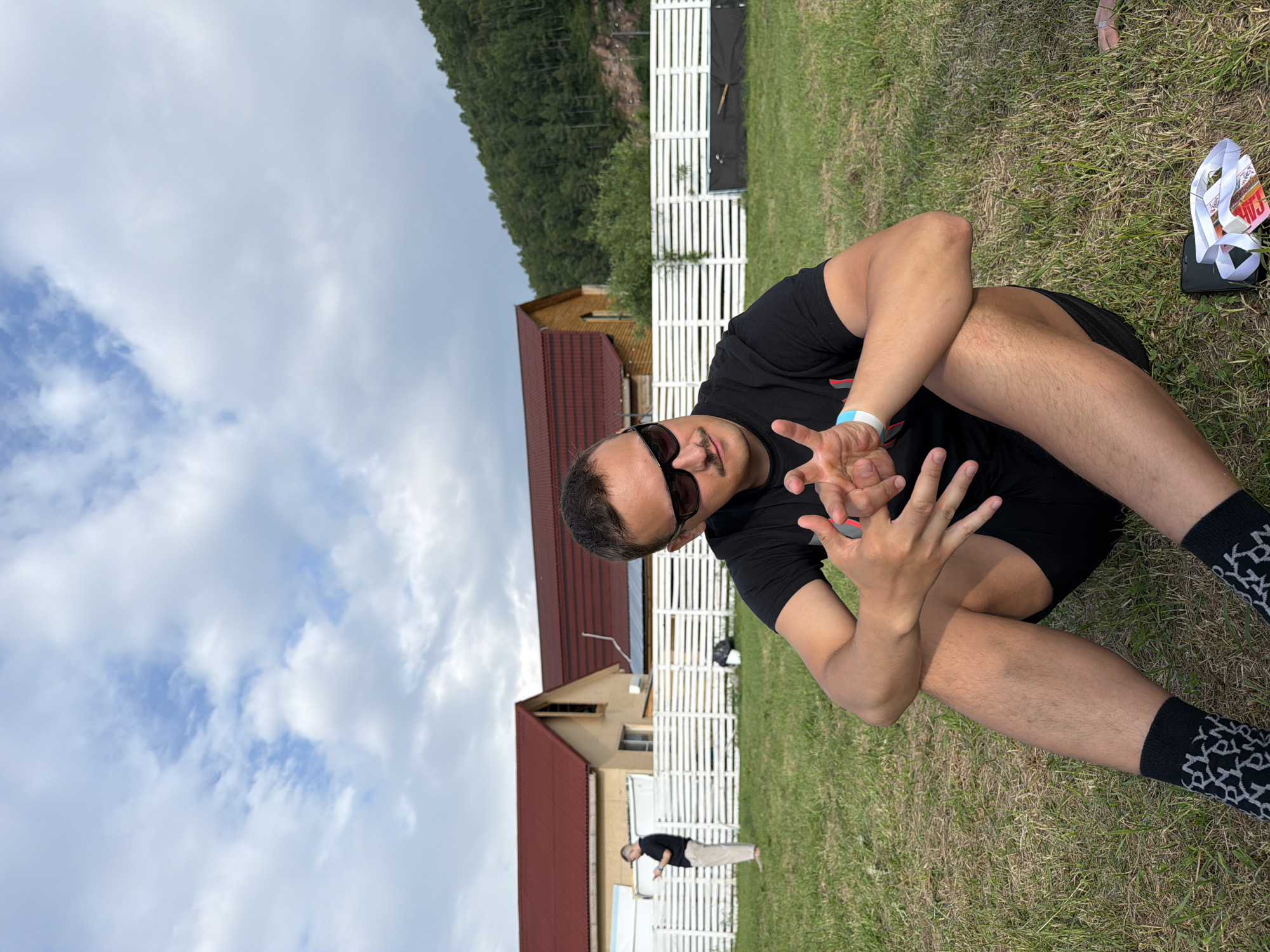 A man in sunglasses and a black t-shirt sits on the grass, facing forward with his legs crossed. He is wearing black shorts and black socks with a white pattern. His hands are raised in front of him, palms open, and he appears to be making a "peace" sign with his right hand. To his right, on the grass, is a black phone with a white lanyard and a colorful piece of paper or card attached to it, possibly a ticket or voucher.
In the background, there is a wooden building with a red roof and another lighter-colored building with a red roof. Behind the buildings, a white wooden fence stretches across the scene. Beyond the fence, a forested hillside rises, and a cloudy sky with patches of blue is visible. The grass appears somewhat dry, suggesting a possibly warm day. Another person in a black shirt and light-colored pants is visible in the distance, standing near the fence.
The scene appears to be outdoors at an event or festival, given the presence of the ticket-like object, the fence, and the general setting. The lighting suggests it is daytime, and the weather is partly cloudy. The man's pose and gesture suggest a relaxed or playful mood. No text is legible in the image, except for the pattern on the man's socks. The location is identified as Дрокино, Russia.